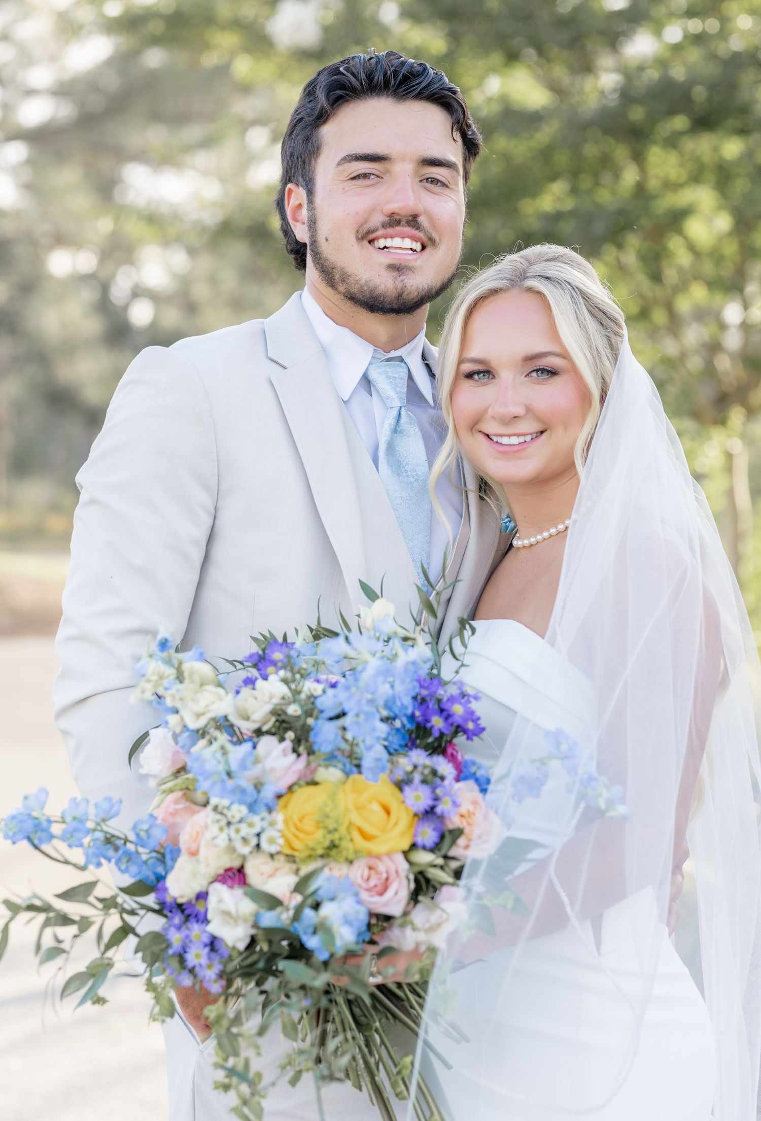 Smiling bride and groom posing outdoors with vibrant blue, yellow, and pink wedding bouquet.