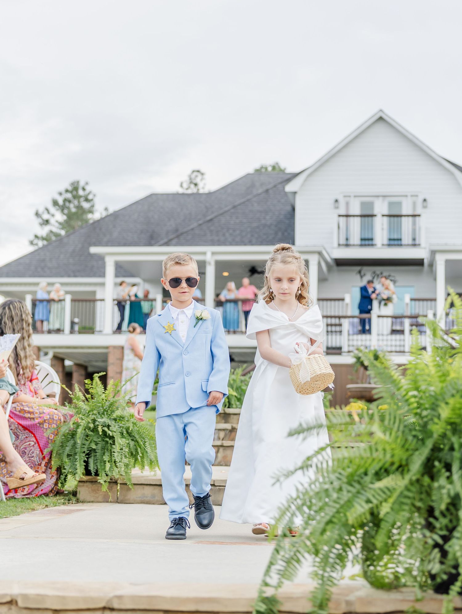Adorable ring bearer in blue suit and flower girl in white dress walk down outdoor wedding aisle.