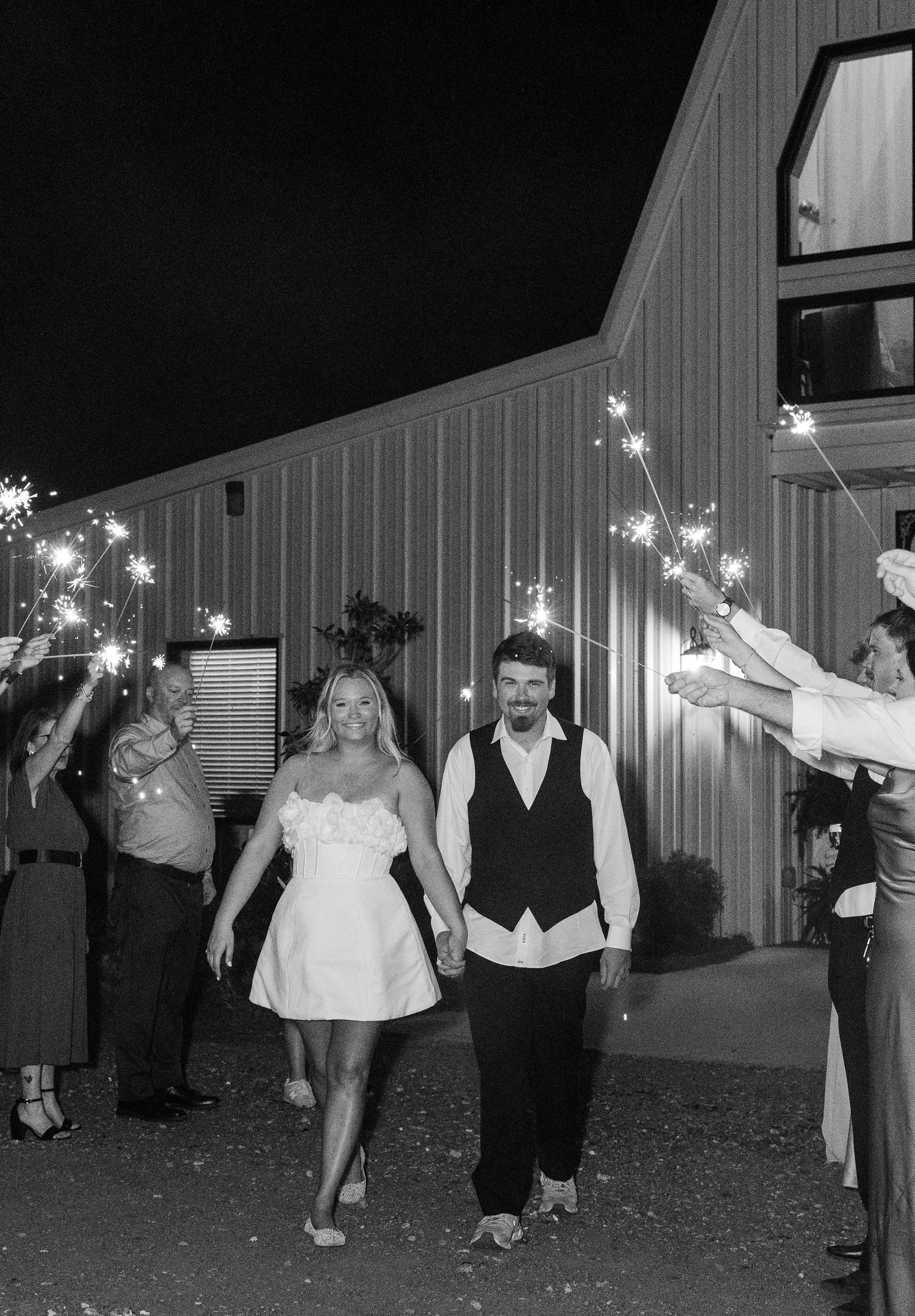 Bride and groom exit wedding venue through sparkler send-off tunnel at night in black and white.