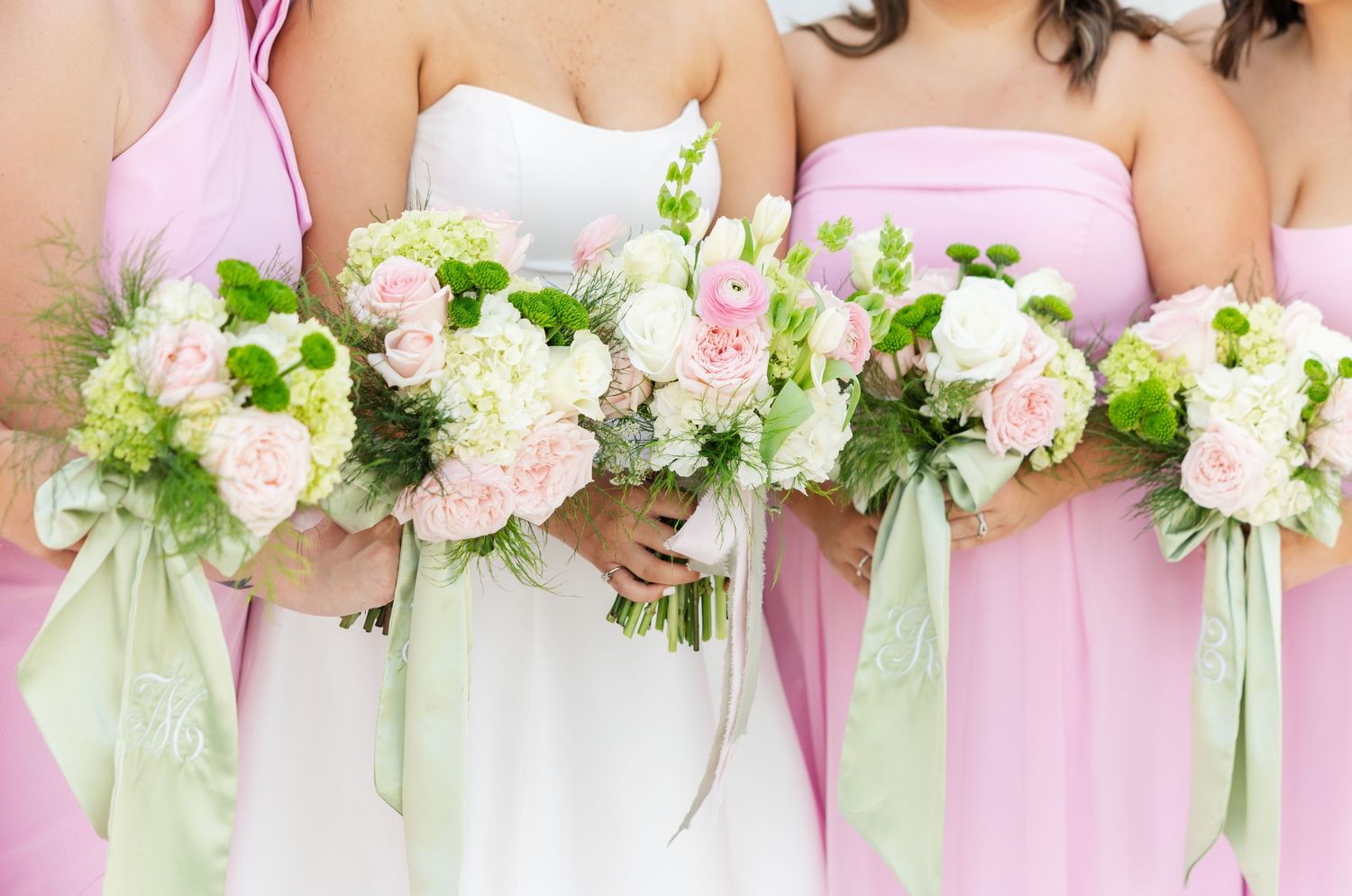 Bridesmaids in pink dresses holding white and pink floral bouquets with green ribbon accents at a wedding.