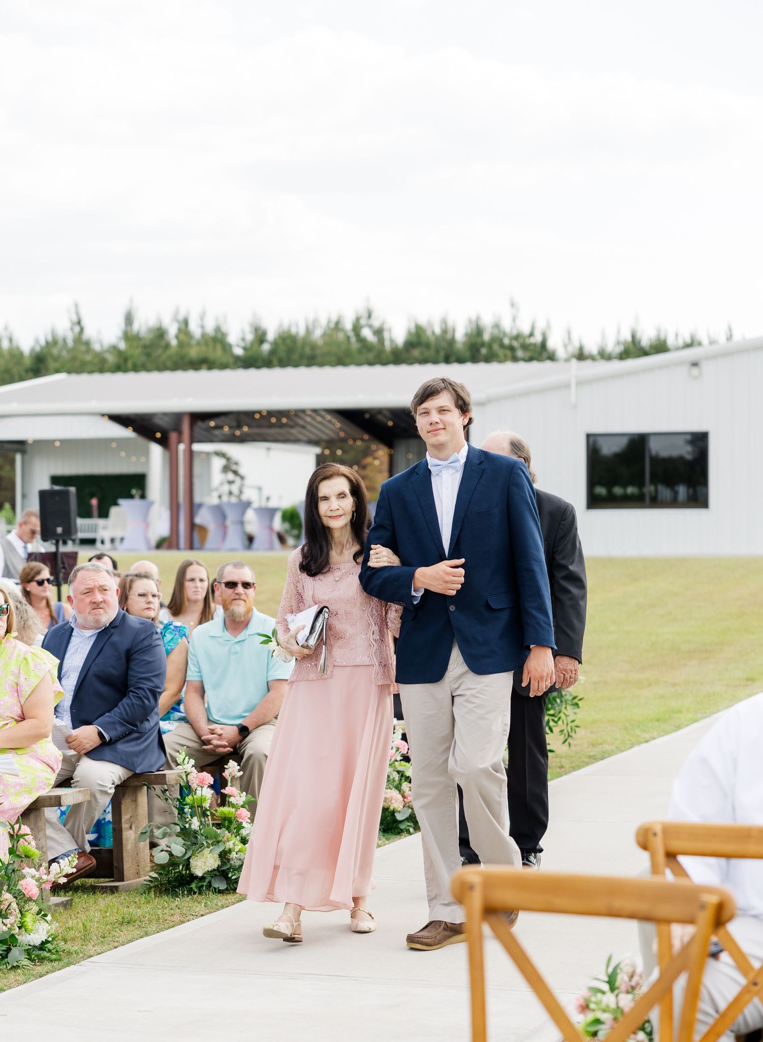 Mother of the bride in pink dress escorted down the outdoor wedding aisle by a groomsman in navy blazer.