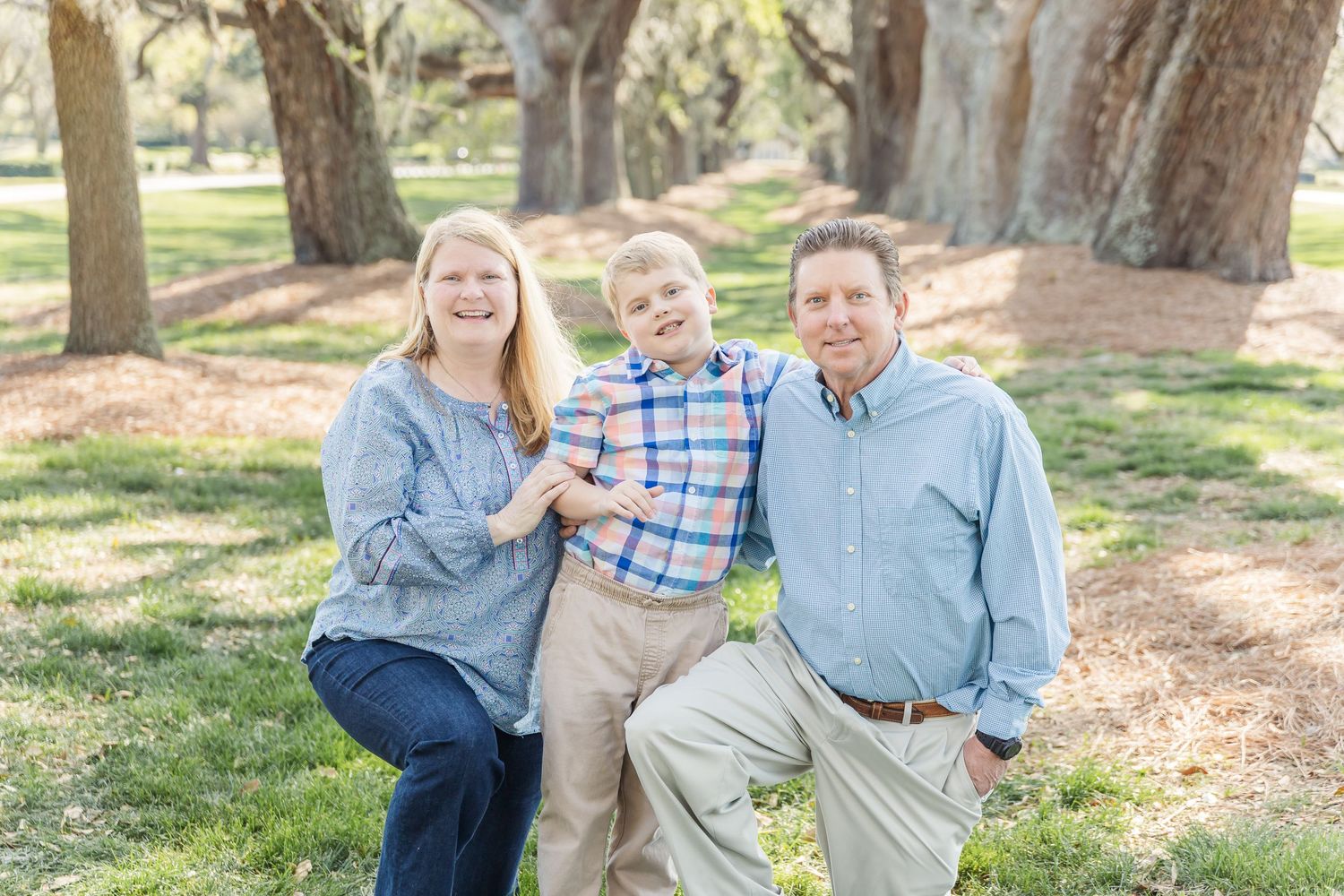 Family of three posing together on grass path under large oak trees.