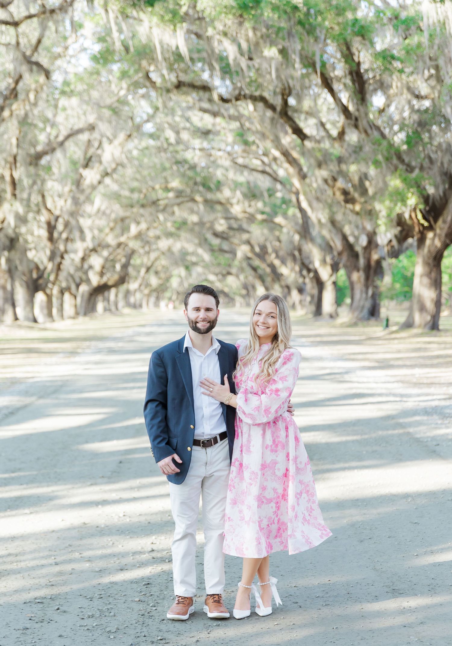Couple poses under canopy of oak trees along scenic dirt road.