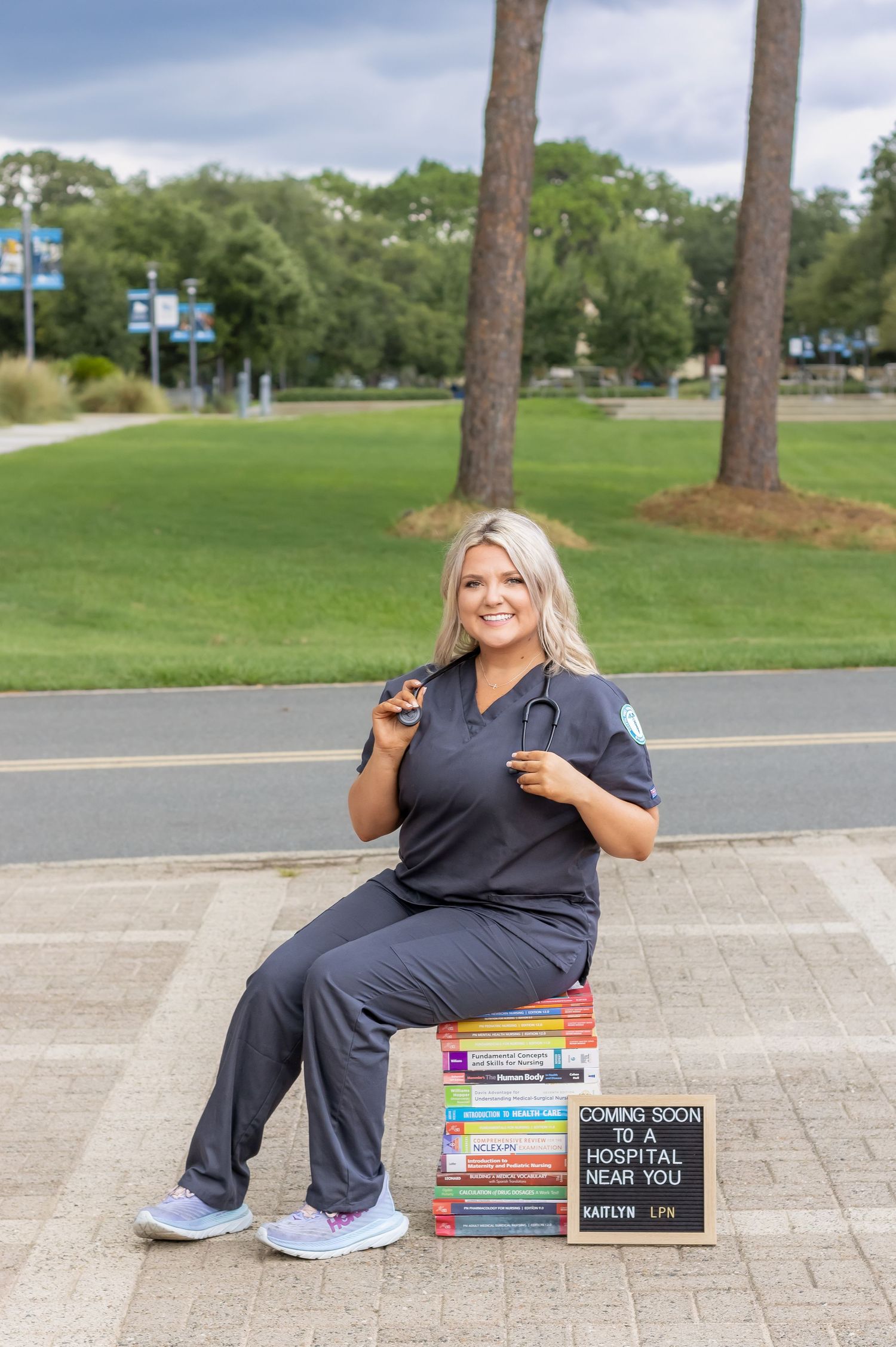 Healthcare worker in scrubs poses with textbooks and graduation sign outdoors on campus.
