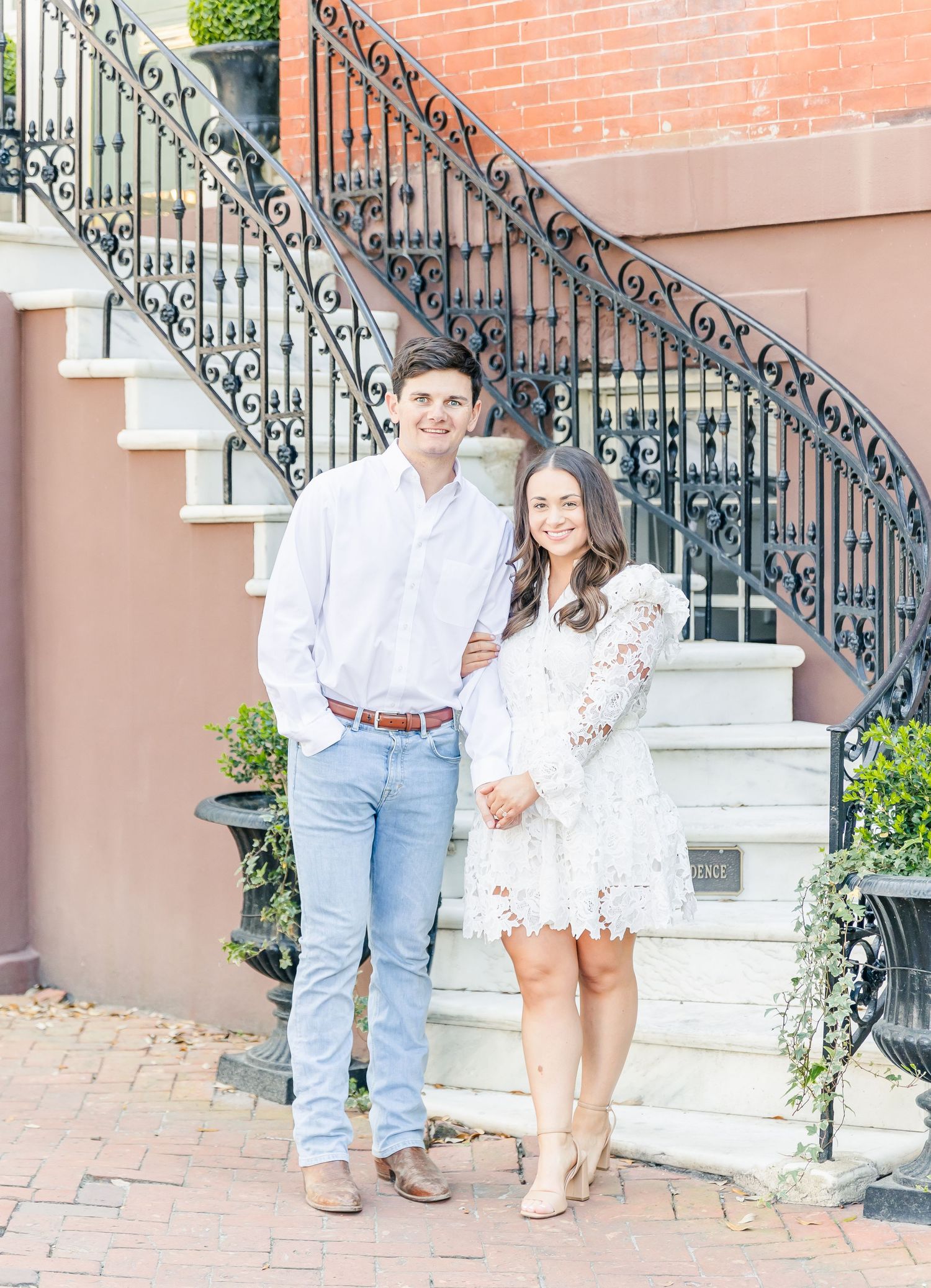 A couple poses on stone steps with ornate wrought iron railings in an urban setting.