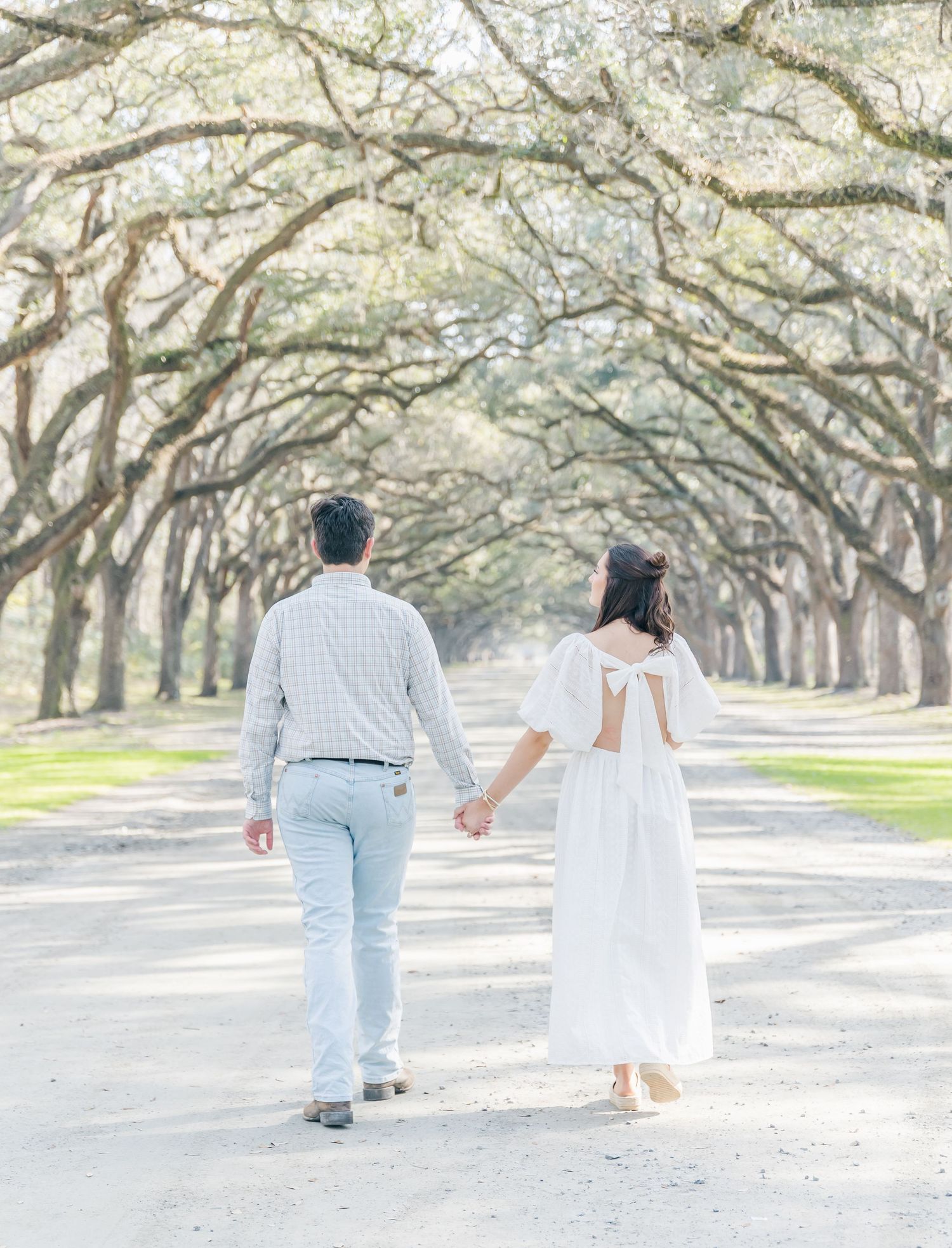 A couple walks away from camera down a scenic tree-lined path in coordinated light-colored outfits.