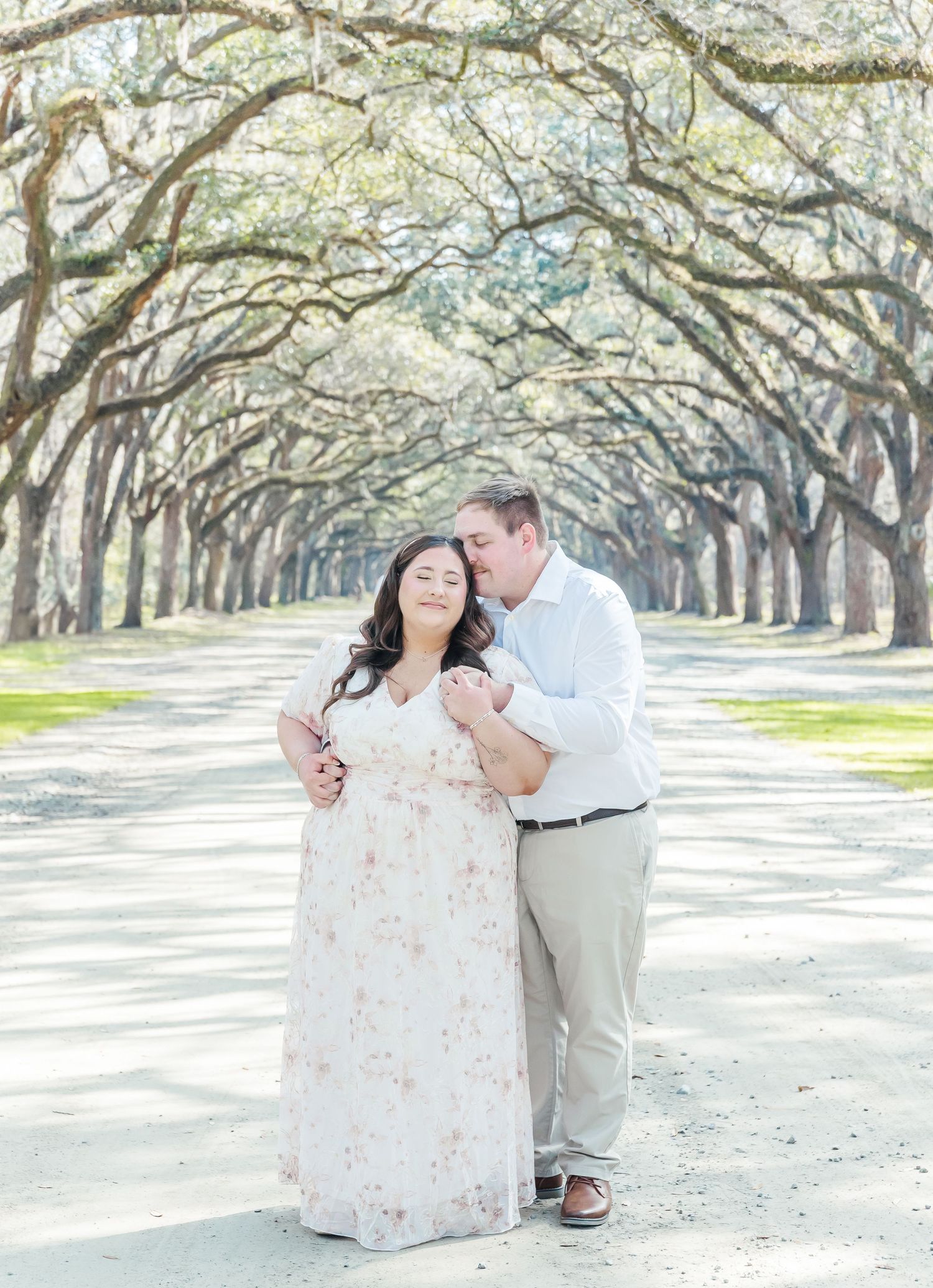 Couple embraces during engagement photos on tree-lined path.