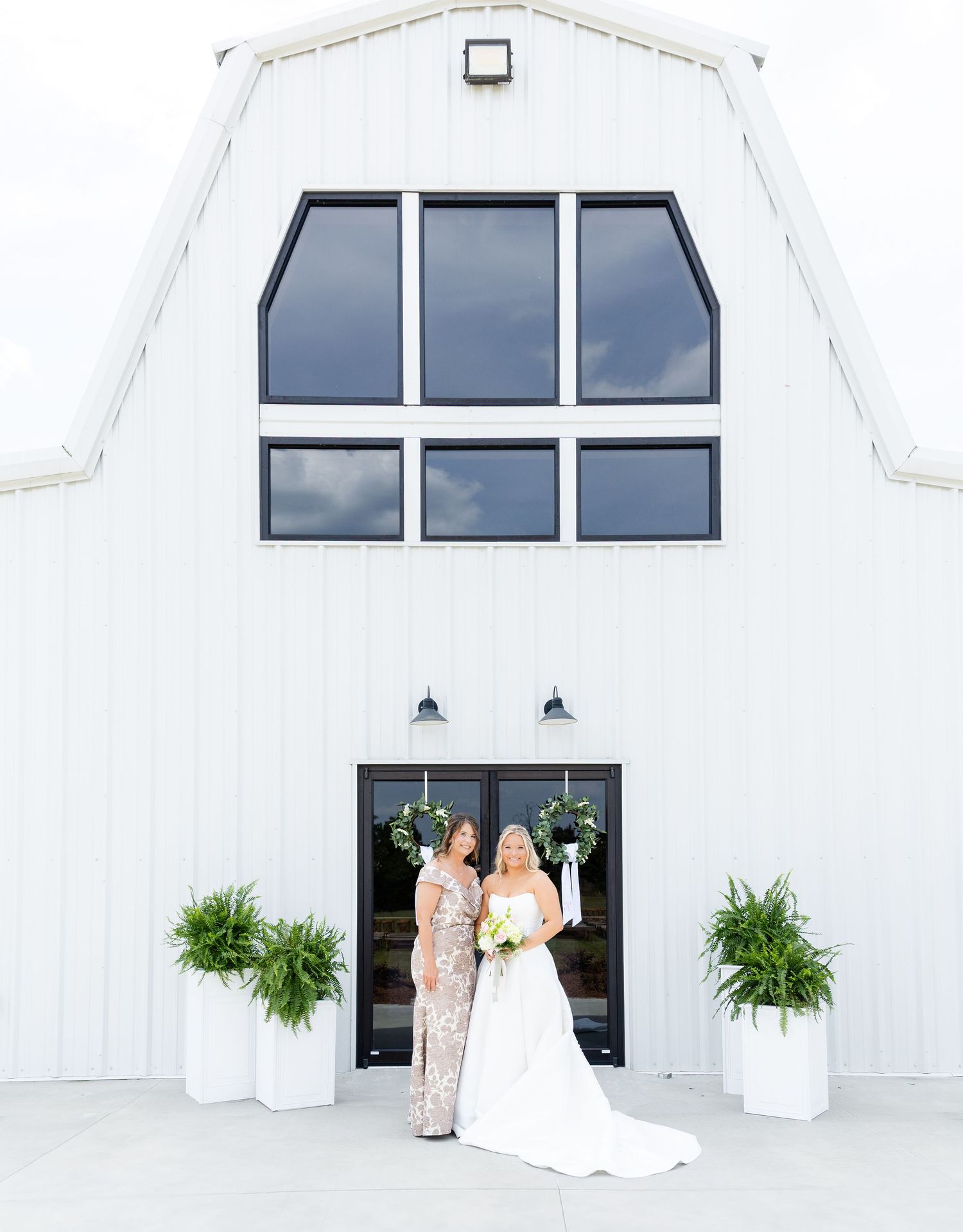 Bride in white gown and mother in sequin dress posing before a white barn wedding venue with fern planters.