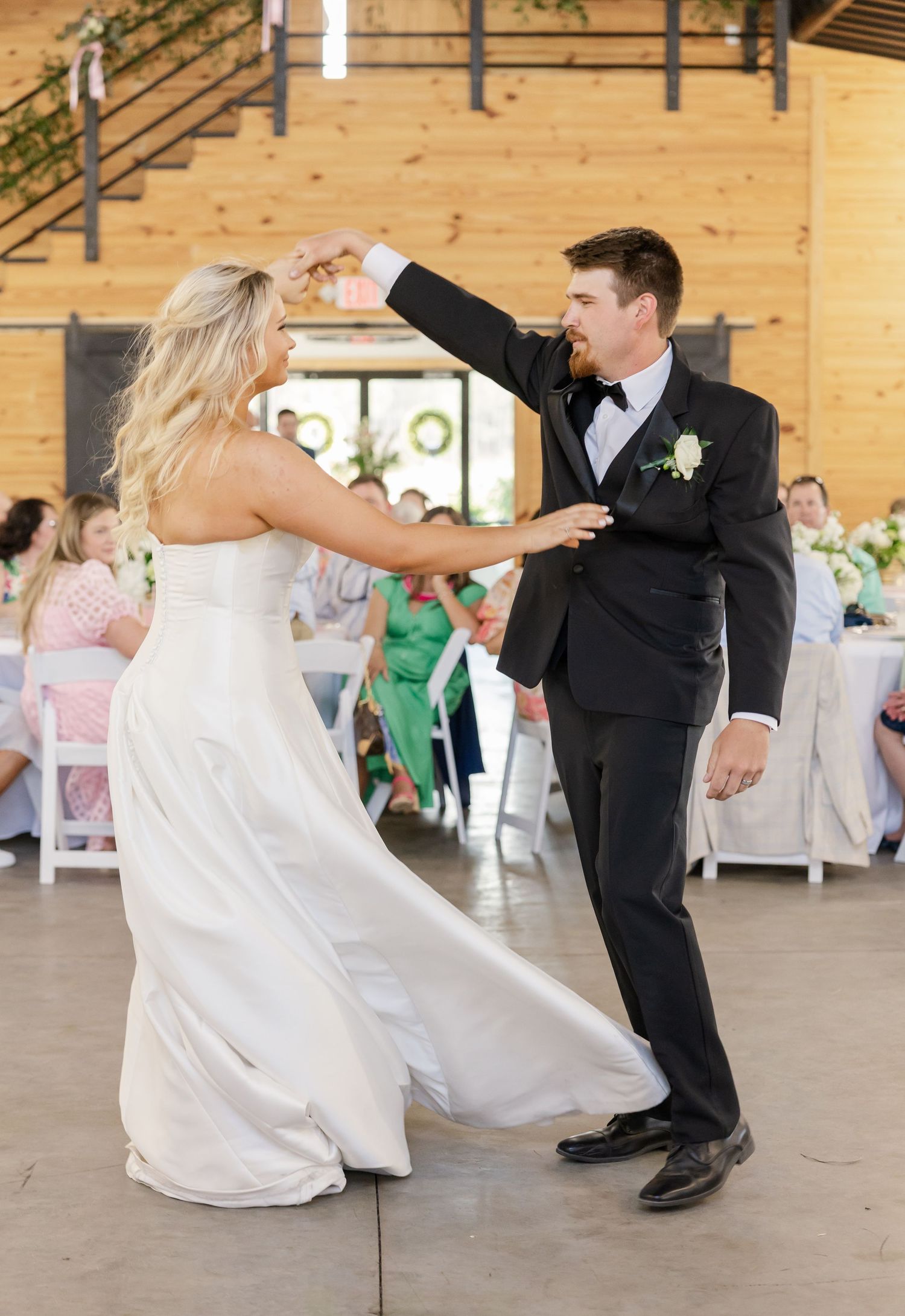 Bride and groom share a joyful first dance in a rustic barn wedding reception venue with guests watching.