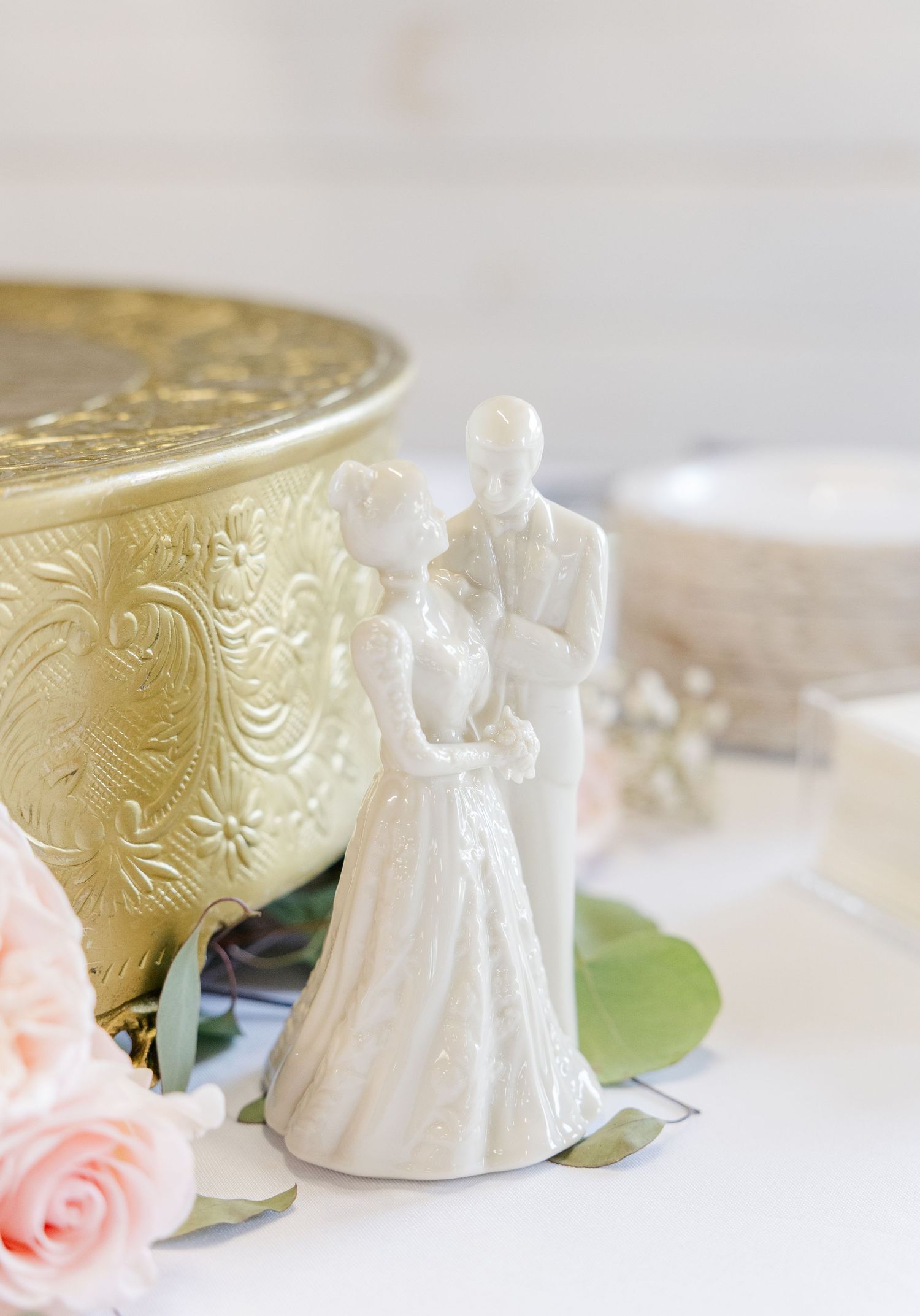 White ceramic bride and groom wedding cake topper figurine beside gold embossed decorative bowl.