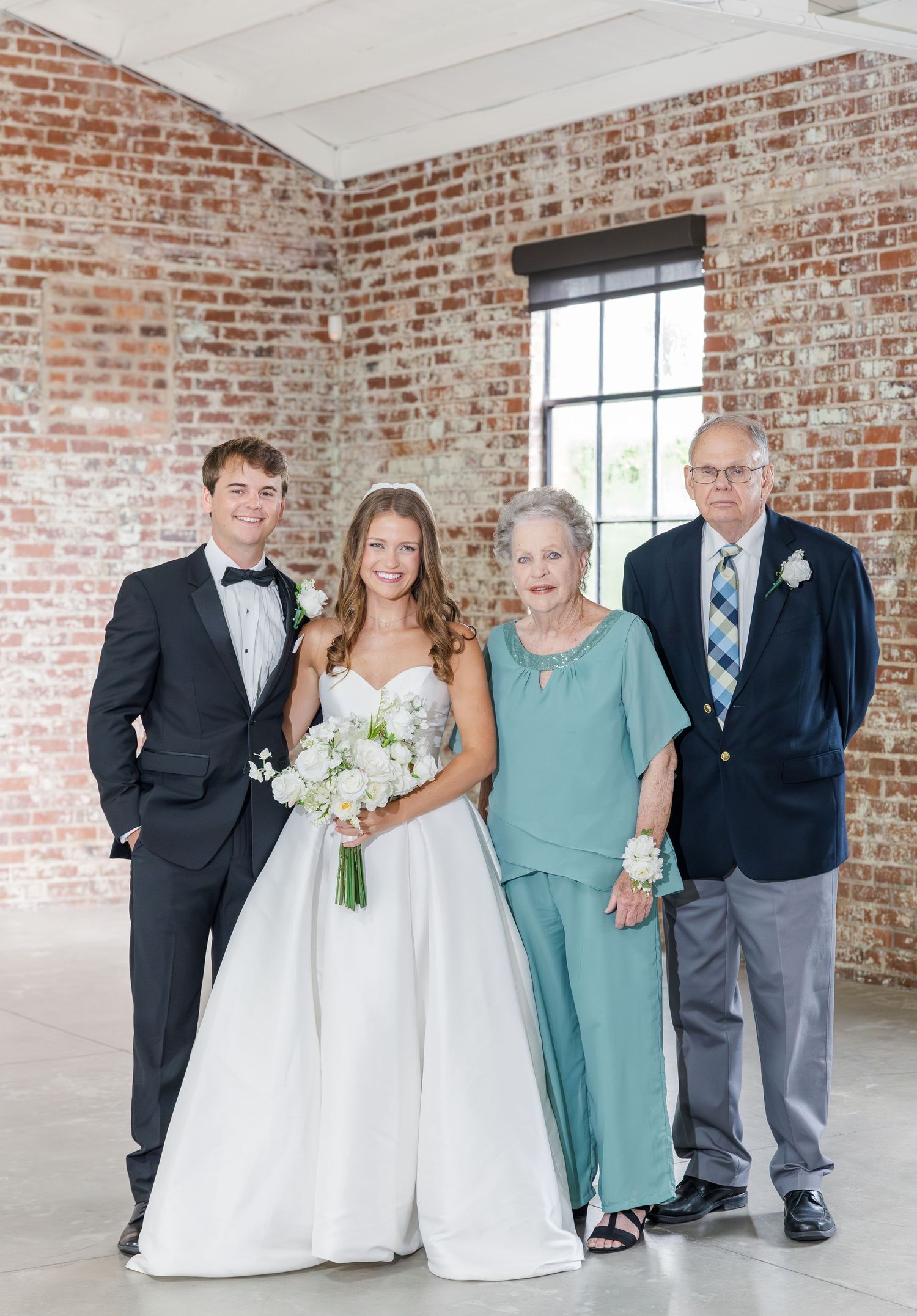 Bride and groom posing with grandparents in front of exposed brick wall at wedding venue.
