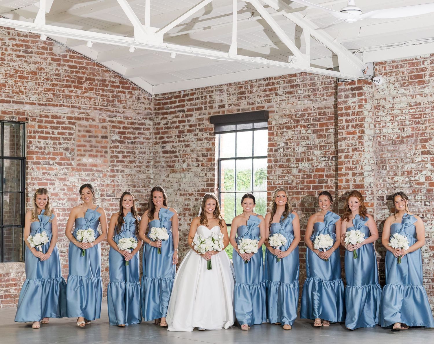 Bride in white ballgown with bridesmaids in dusty blue dresses holding white bouquets in brick venue.