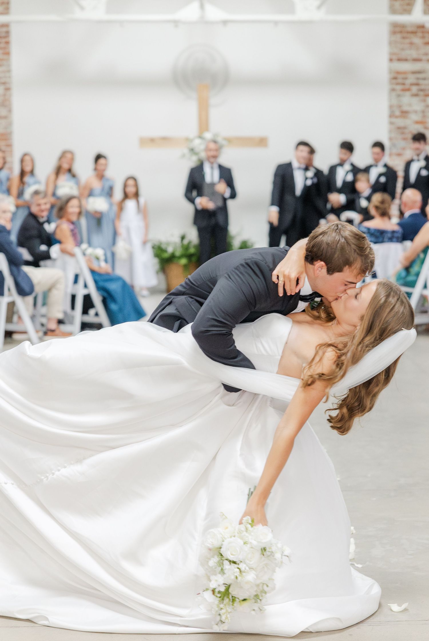 Groom dips bride for a kiss after ceremony in bright brick-walled wedding venue.