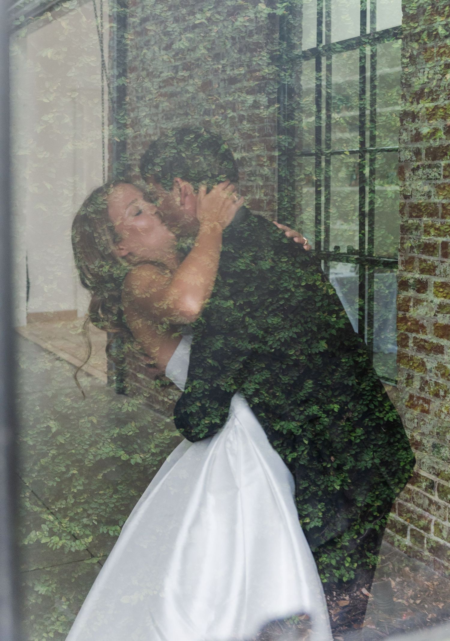 Bride and groom share a romantic kiss through a rain-streaked window with ivy reflections on their wedding day.