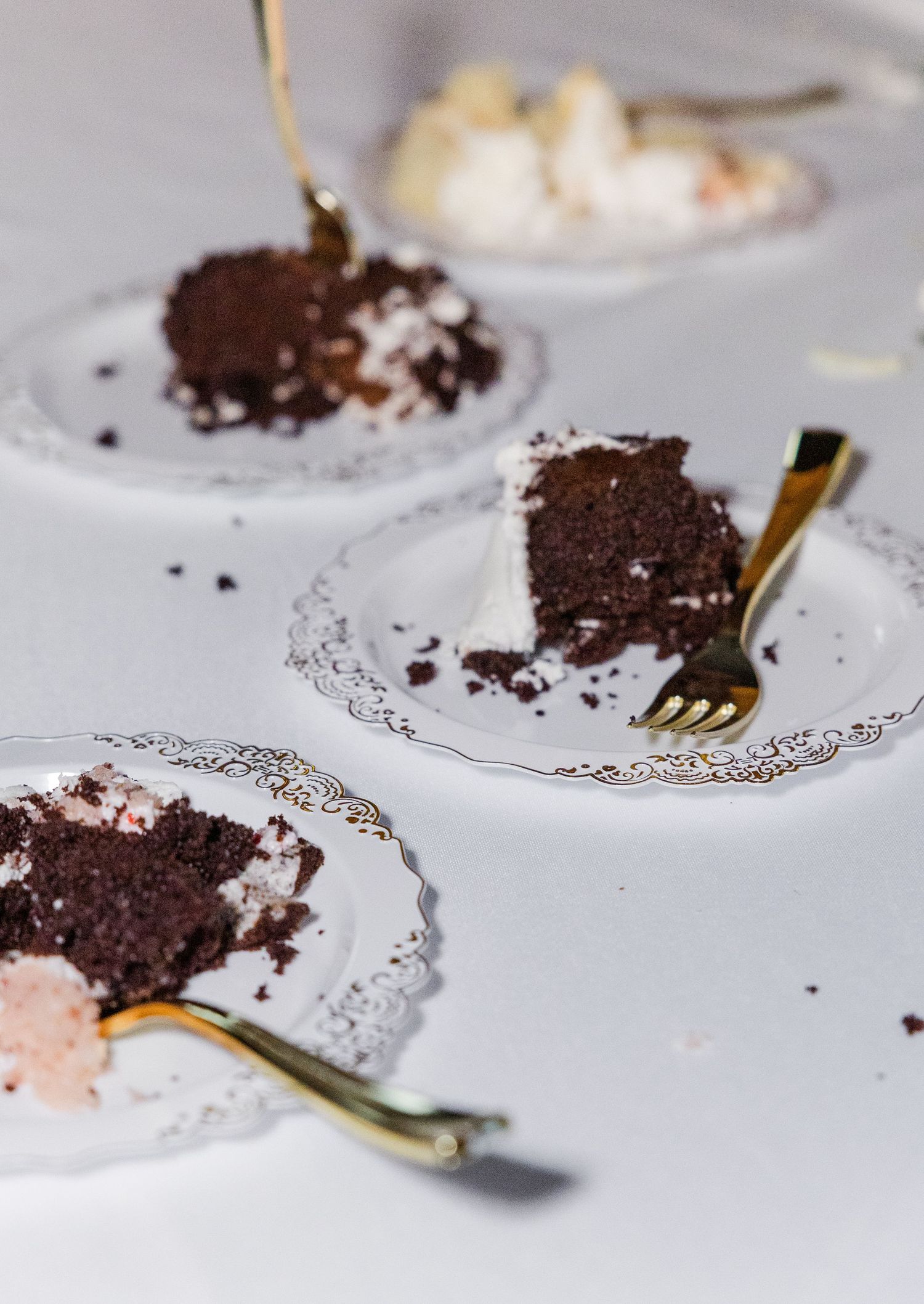 Three plates of chocolate wedding cake slices with white frosting and gold forks on white table.