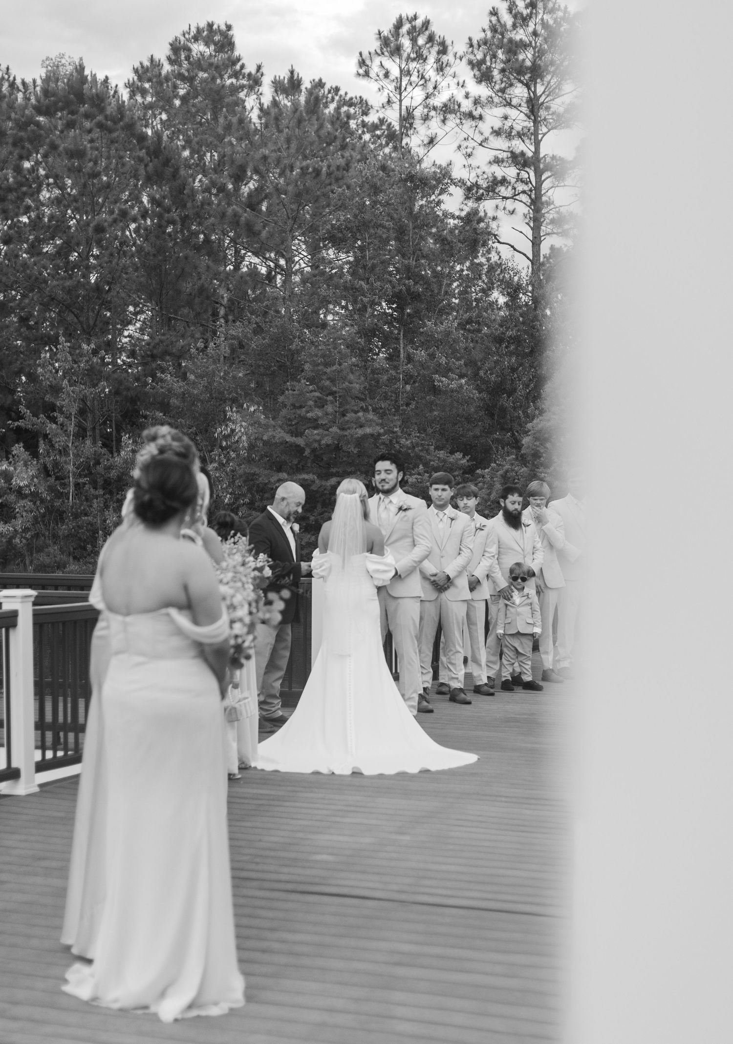Black and white photo of bride walking down aisle toward groom and groomsmen outdoors.