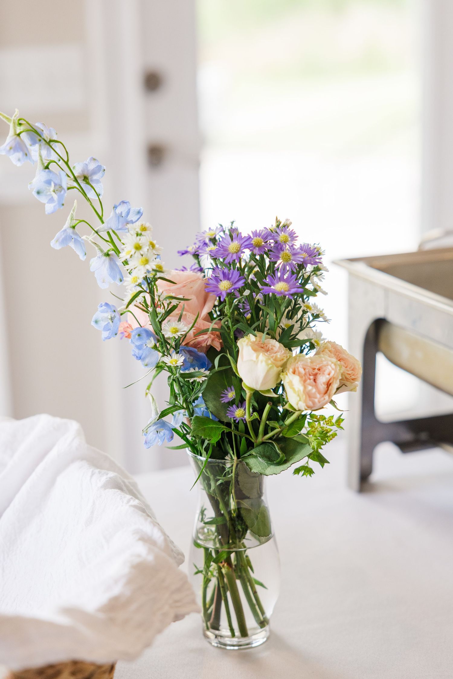 A glass vase of mixed wildflowers including blue bells, purple asters, and peach roses on a white table.