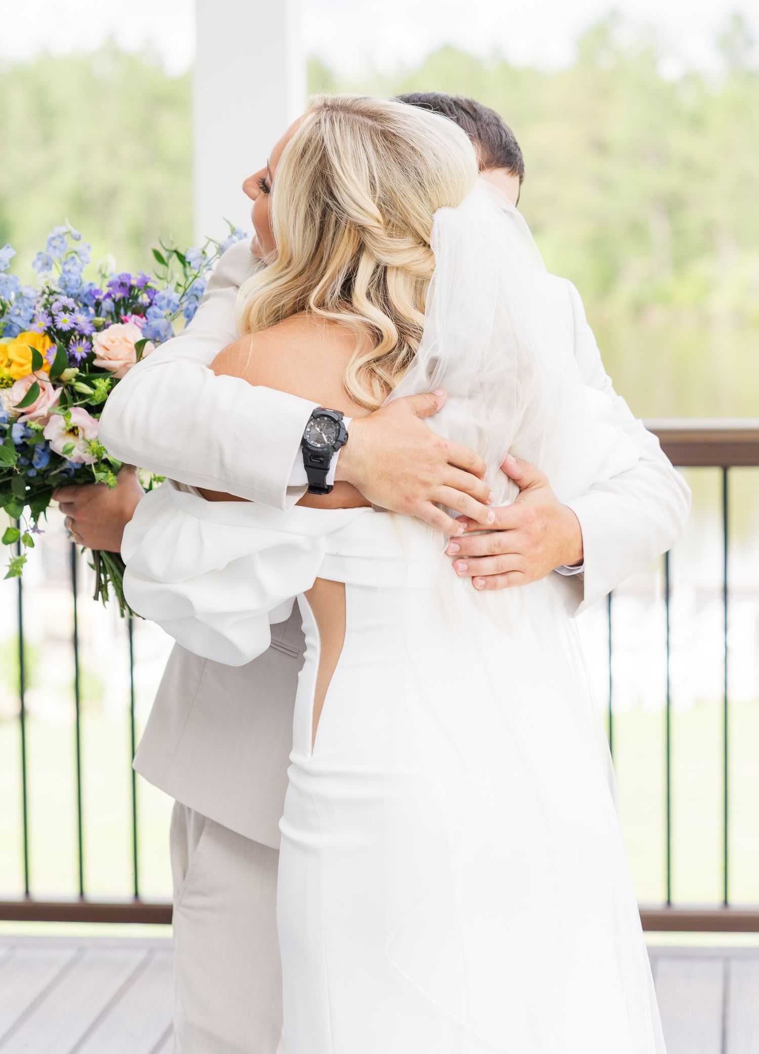 Bride and groom sharing first look embrace on outdoor deck, bride holding colorful wildflower bouquet.