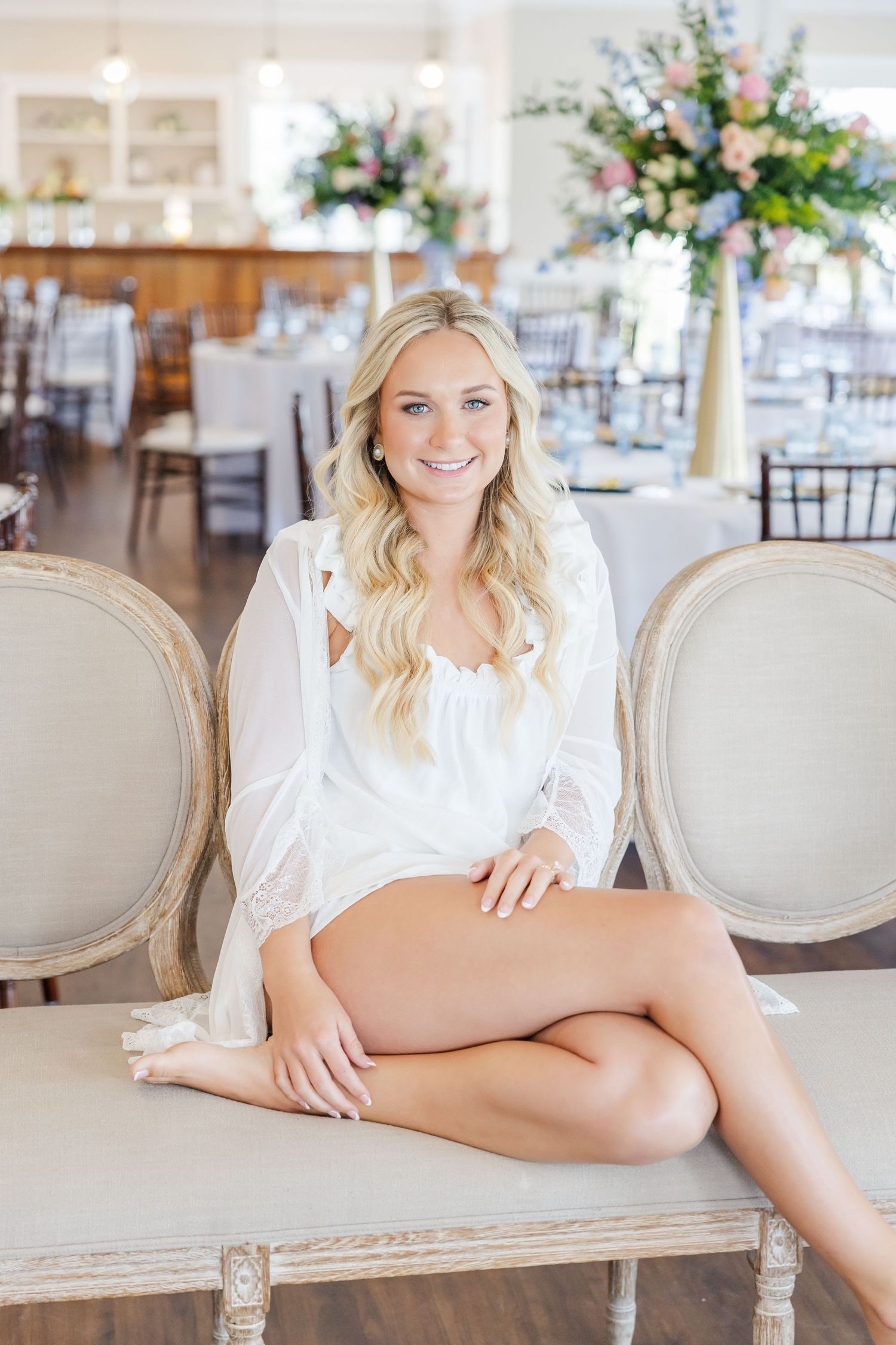 Smiling blonde bride in white lace robe seated on elegant chair in bright wedding venue.
