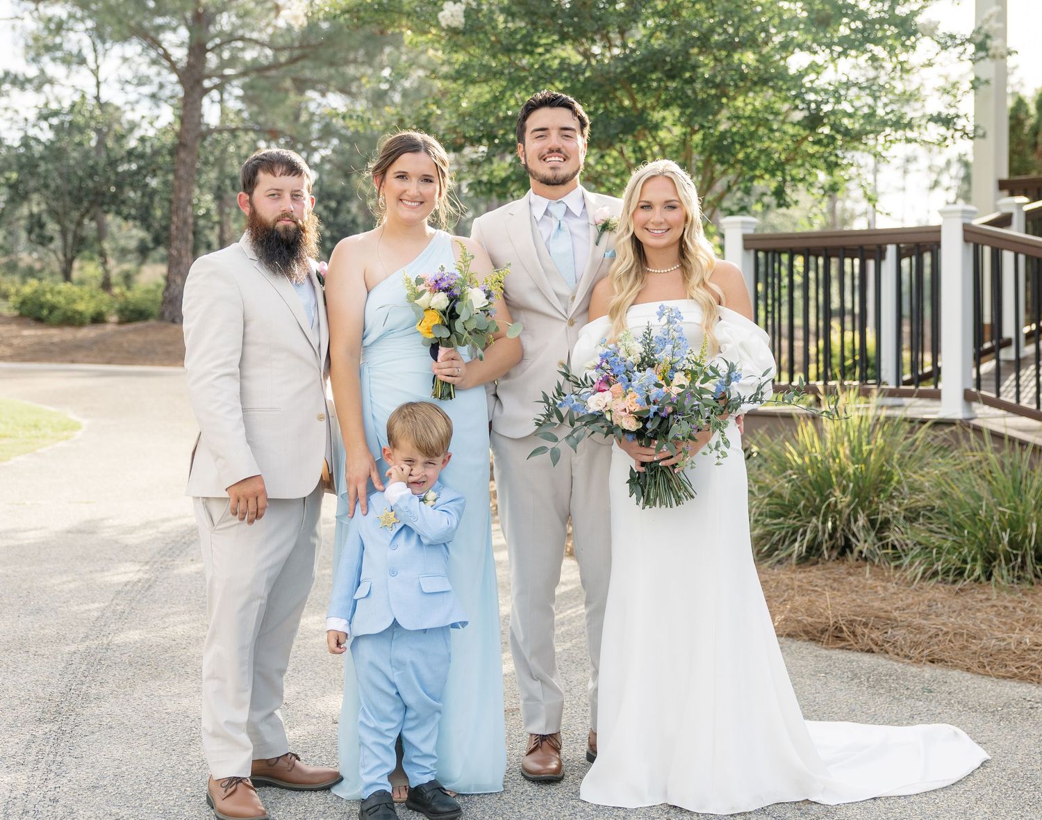 Wedding party group photo with bride, groom, bridesmaid, groomsman, and ring bearer in outdoor venue setting.