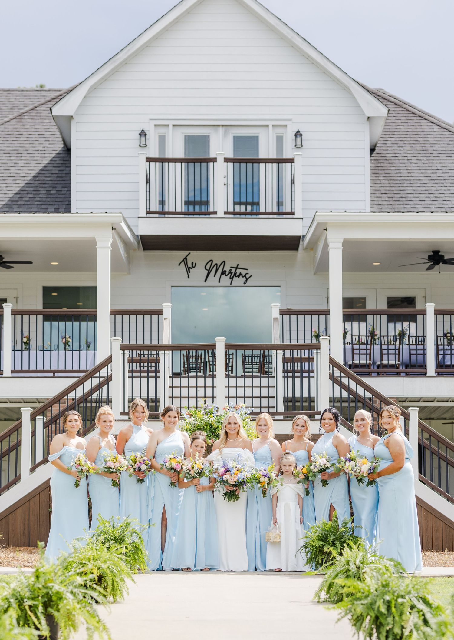 Bride with bridesmaids in light blue gowns posing in front of white farmhouse wedding venue.