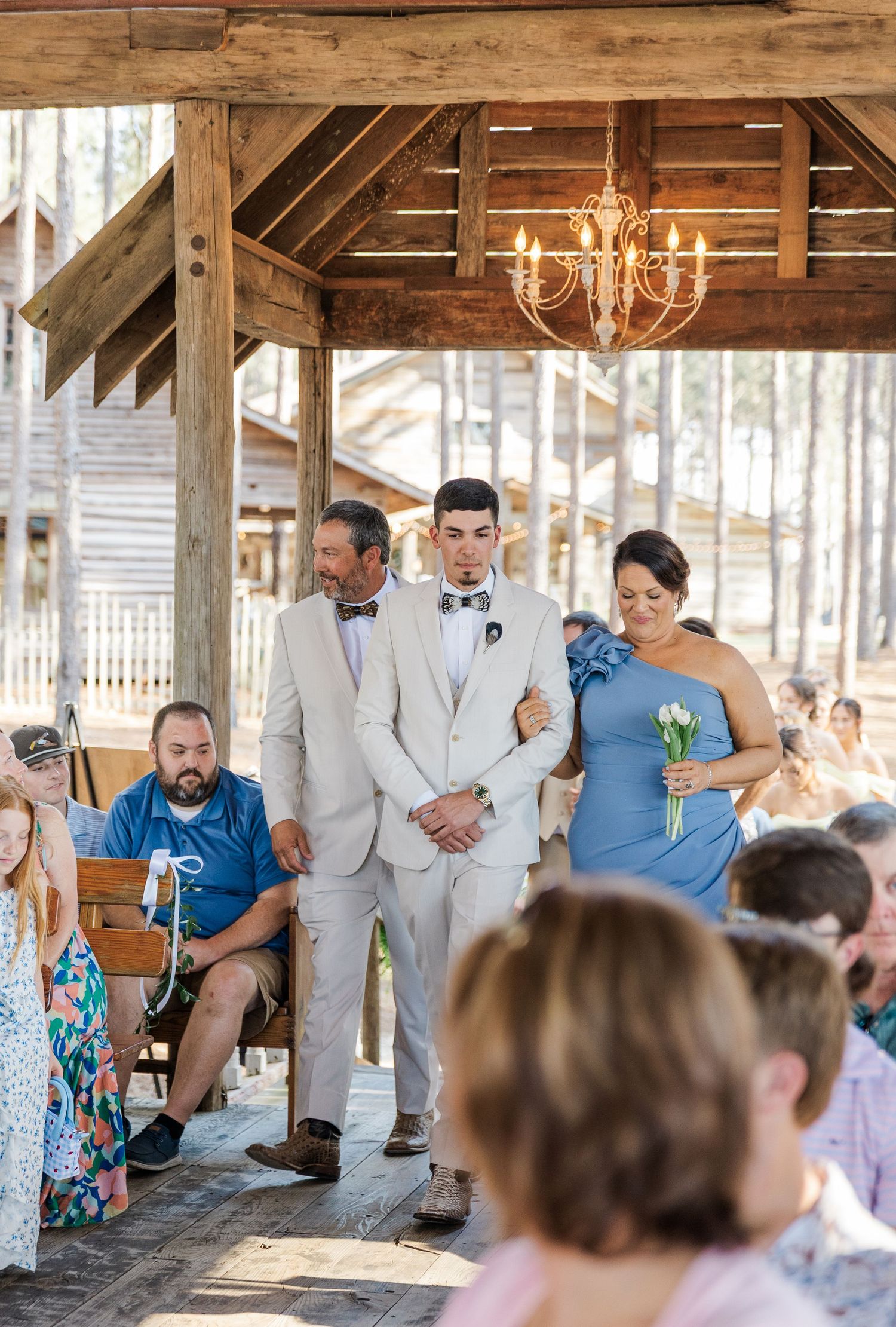 Groom walking down aisle escorted by parents at rustic outdoor wedding ceremony pavilion.