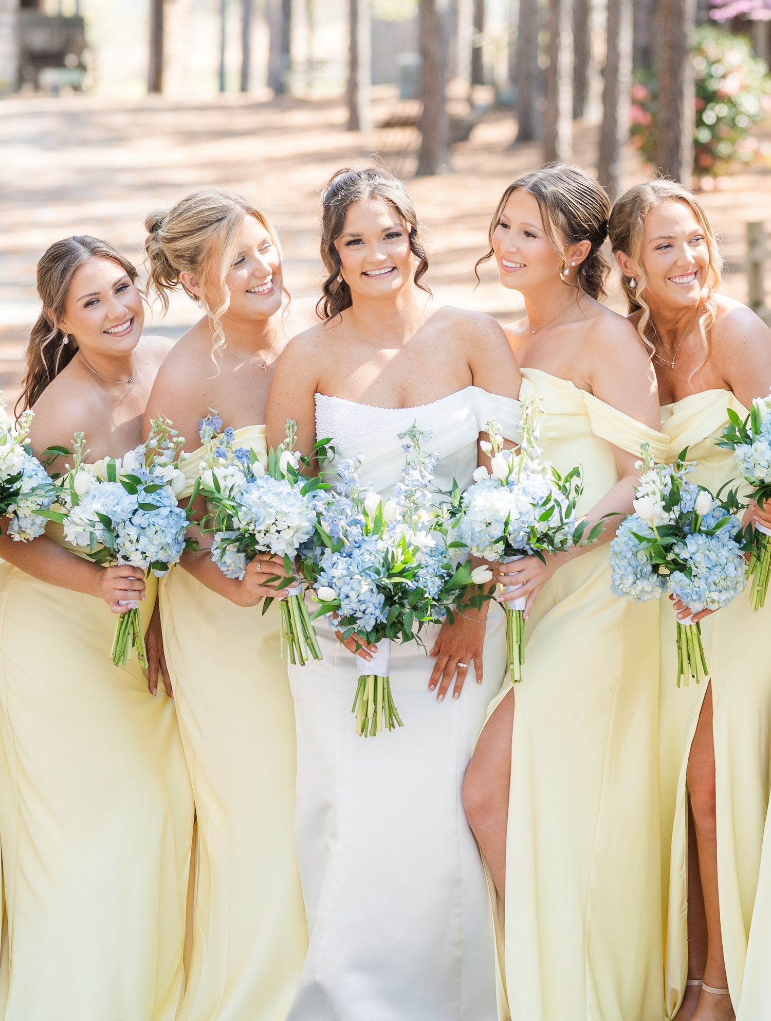Bride in white strapless gown with four bridesmaids in yellow dresses holding blue hydrangea bouquets outdoors.