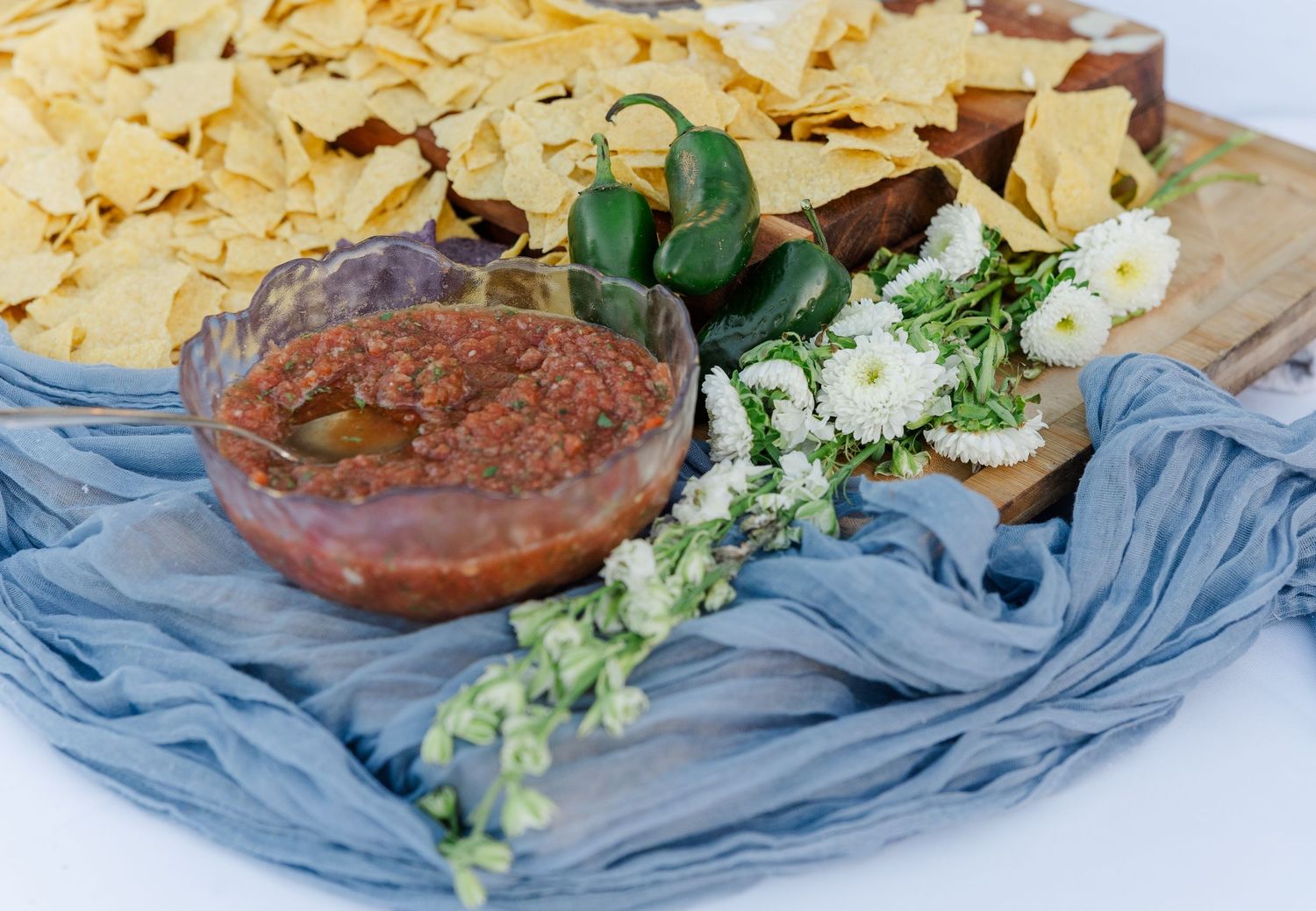 Chips and salsa appetizer display with jalapenos and white flowers on blue fabric.