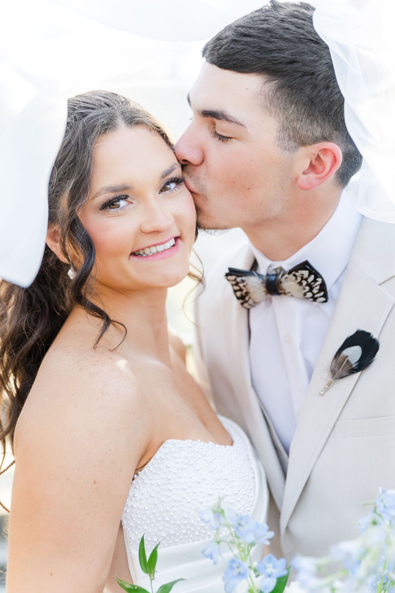 Groom in beige suit kissing smiling bride on the cheek with blue flowers in foreground.