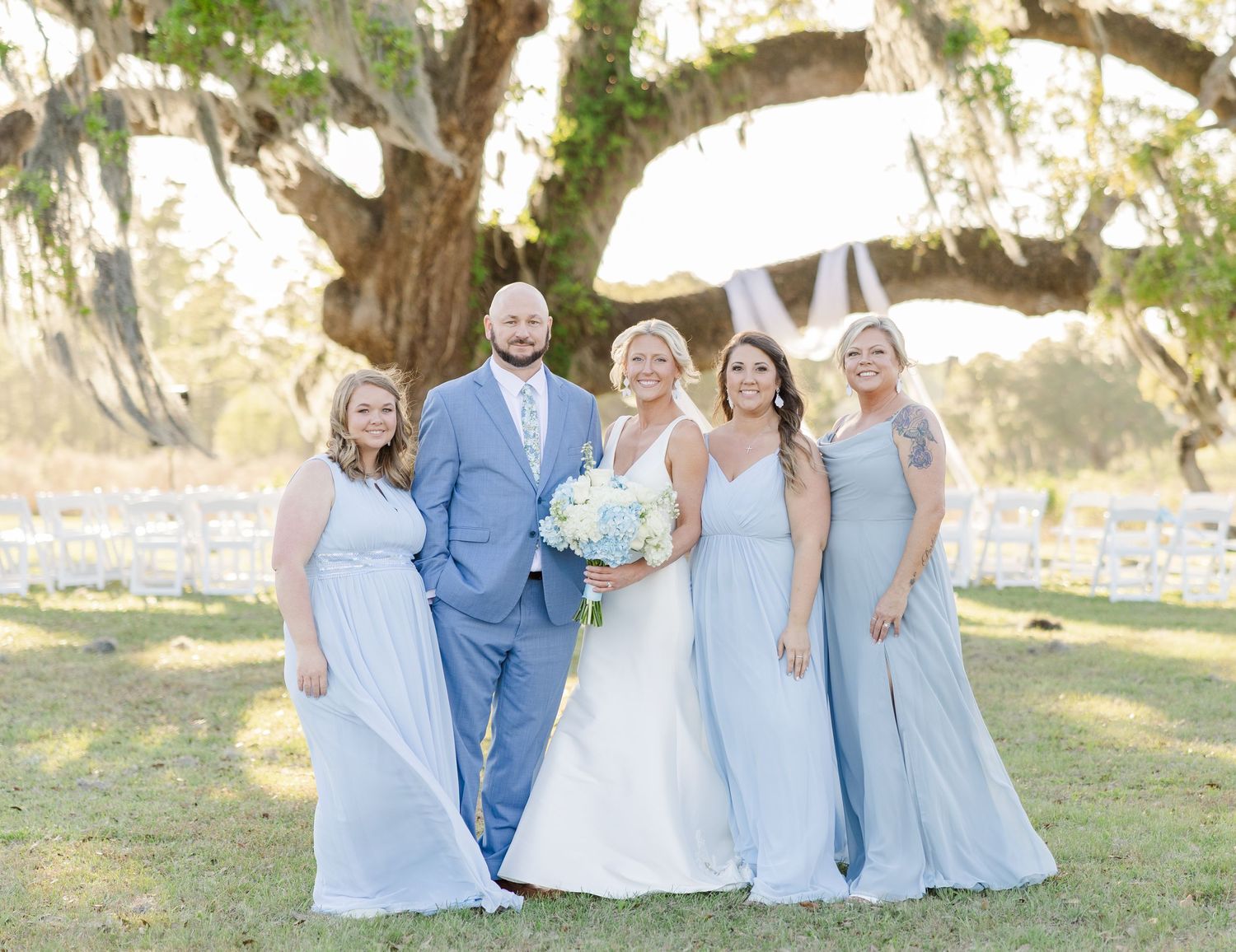 Wedding party in light blue attire posing under large oak tree outdoors.