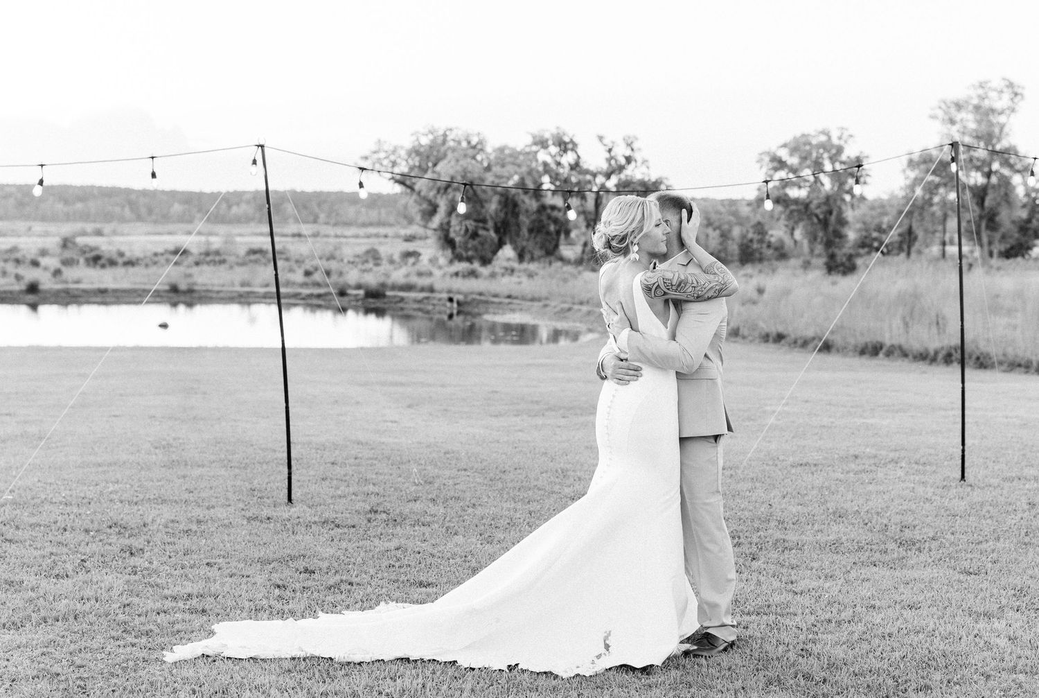 Black and white photo of bride and groom embracing near a pond at their outdoor wedding reception.
