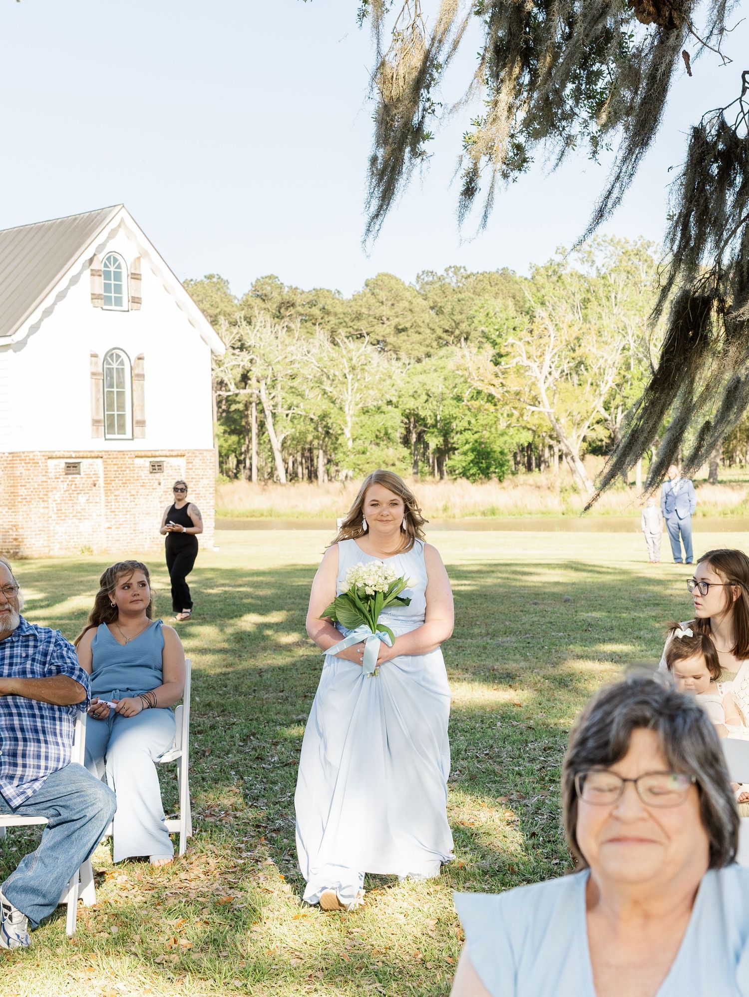 Bridesmaid walks down outdoor aisle carrying white bouquet at sunny garden wedding ceremony.
