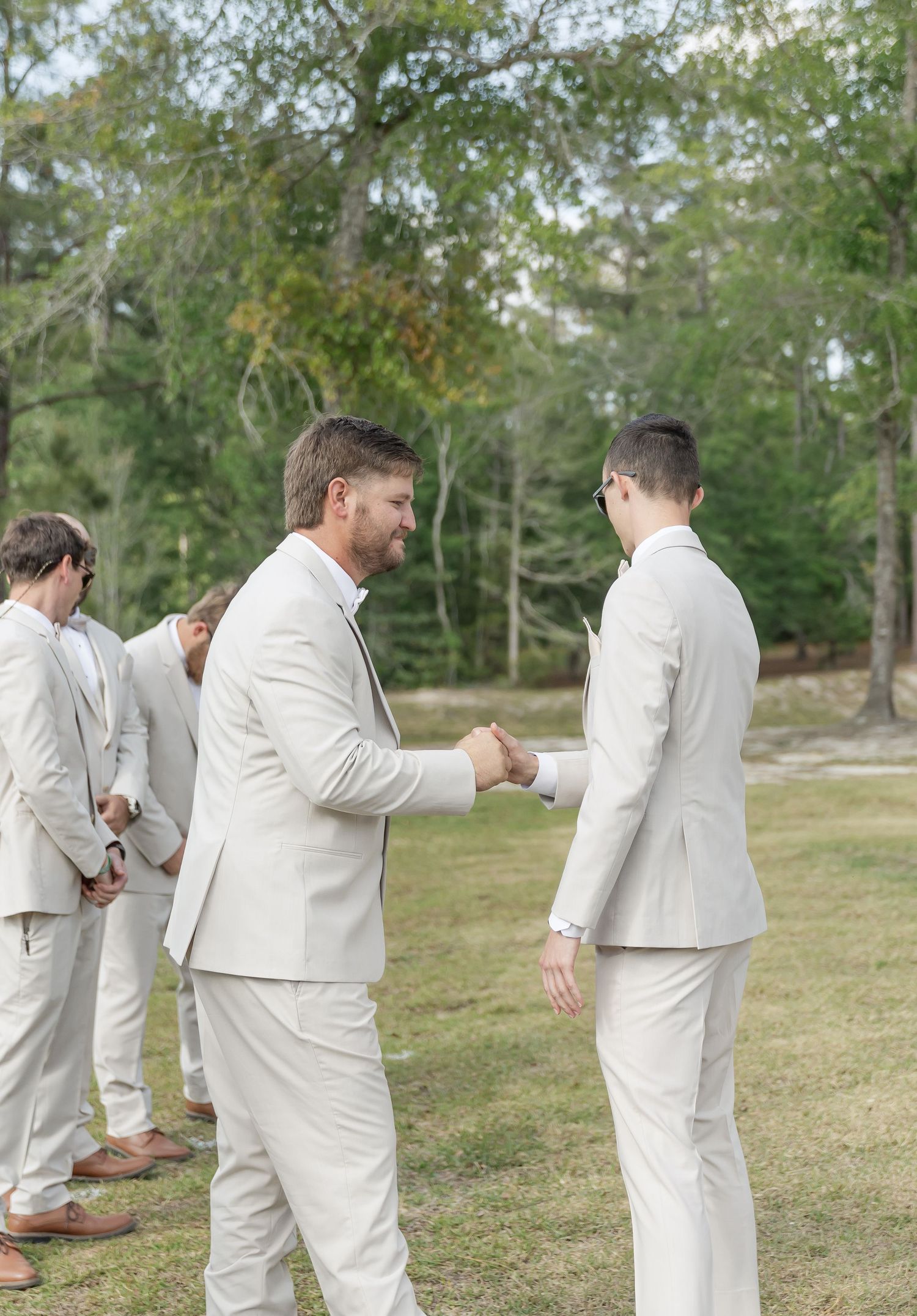 Groomsmen in light colored suits exchange handshakes at outdoor ceremony.