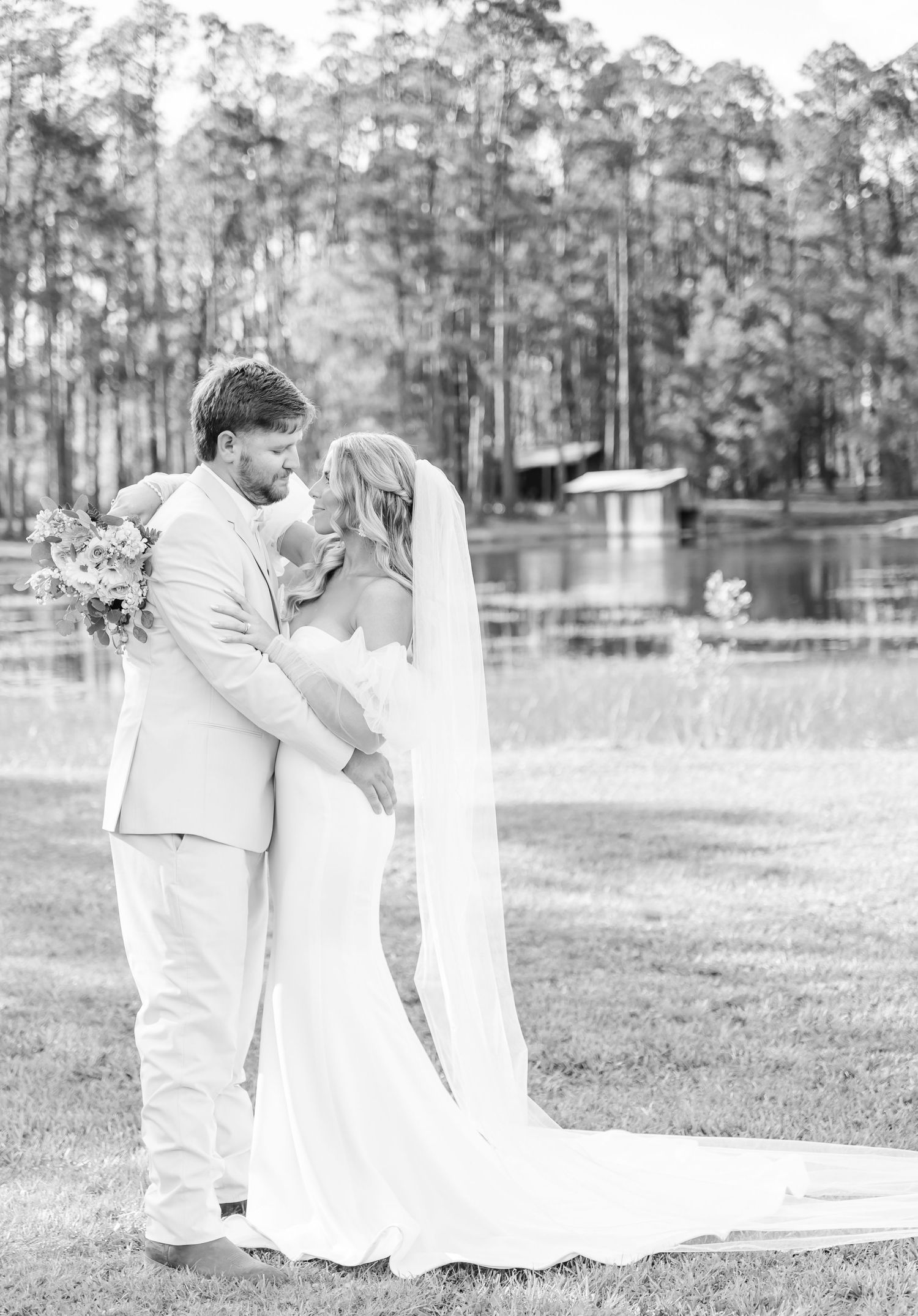 A black and white wedding photo of a couple embracing by a serene lake.
