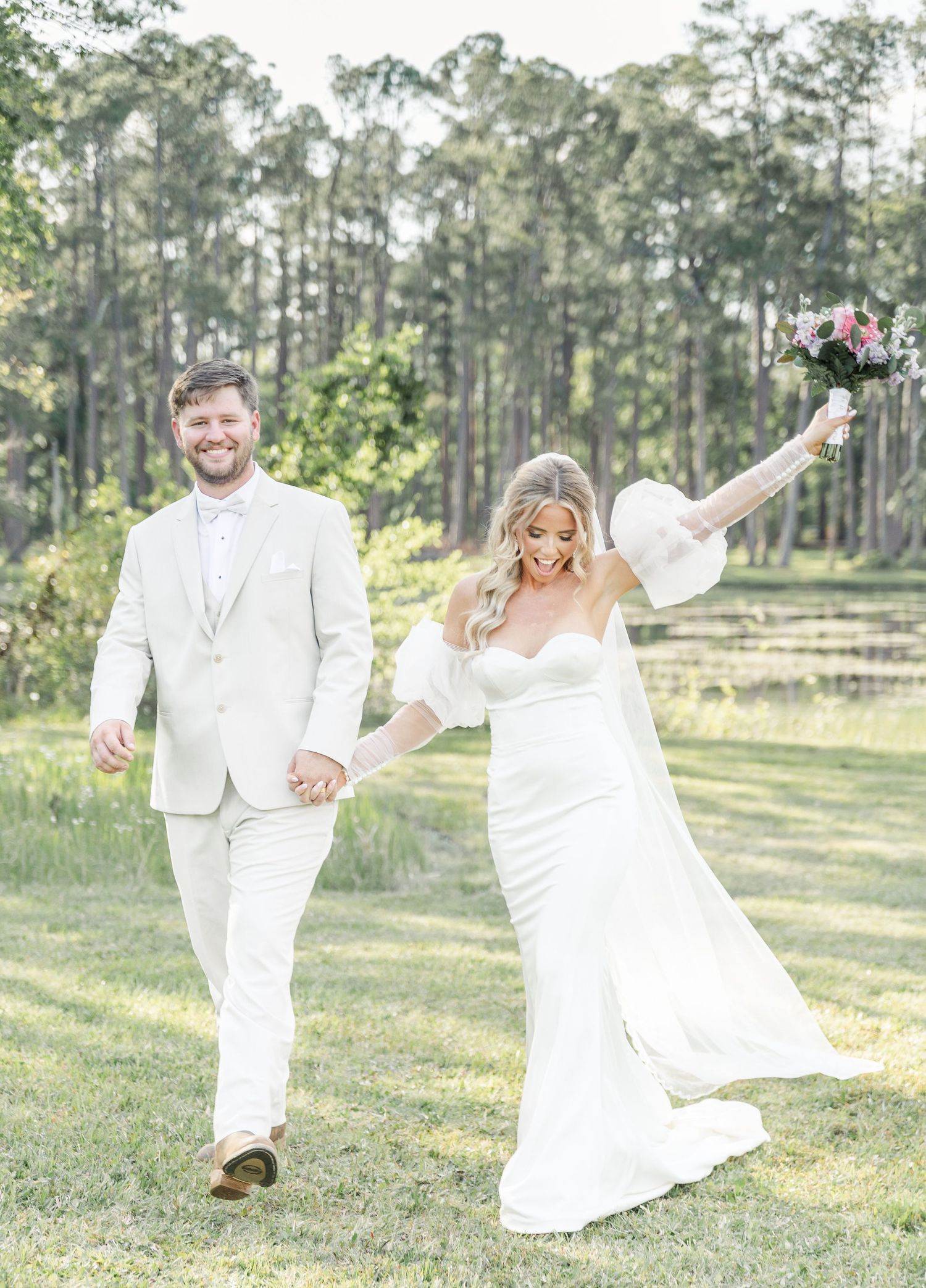 Happy newlyweds in white attire walking together on grass with tall pine trees in background.
