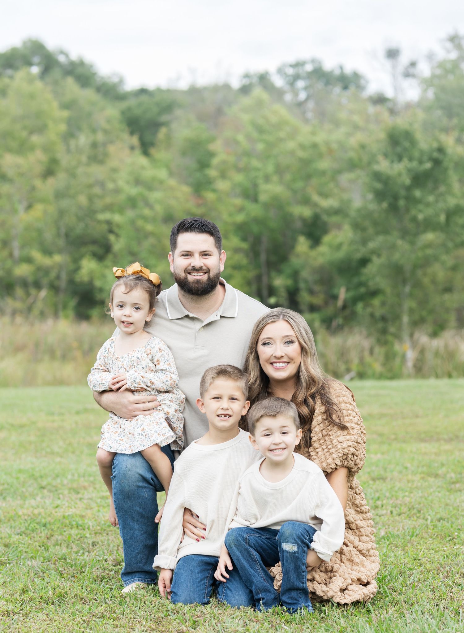 A family wearing coordinated cream and brown outfits poses together on grass during an outdoor photo session.