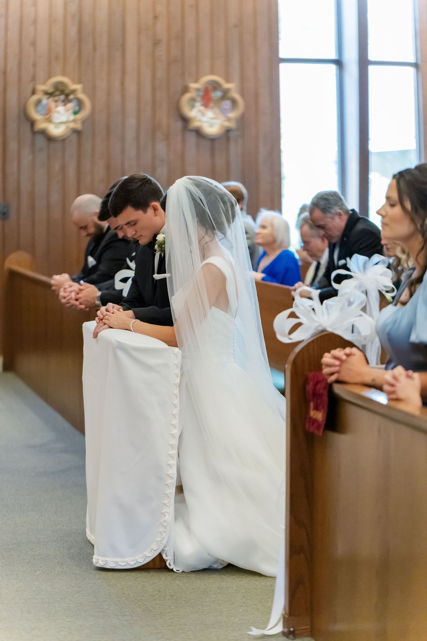 Couples sit in wooden church pews during a wedding ceremony with white veils visible in the foreground.