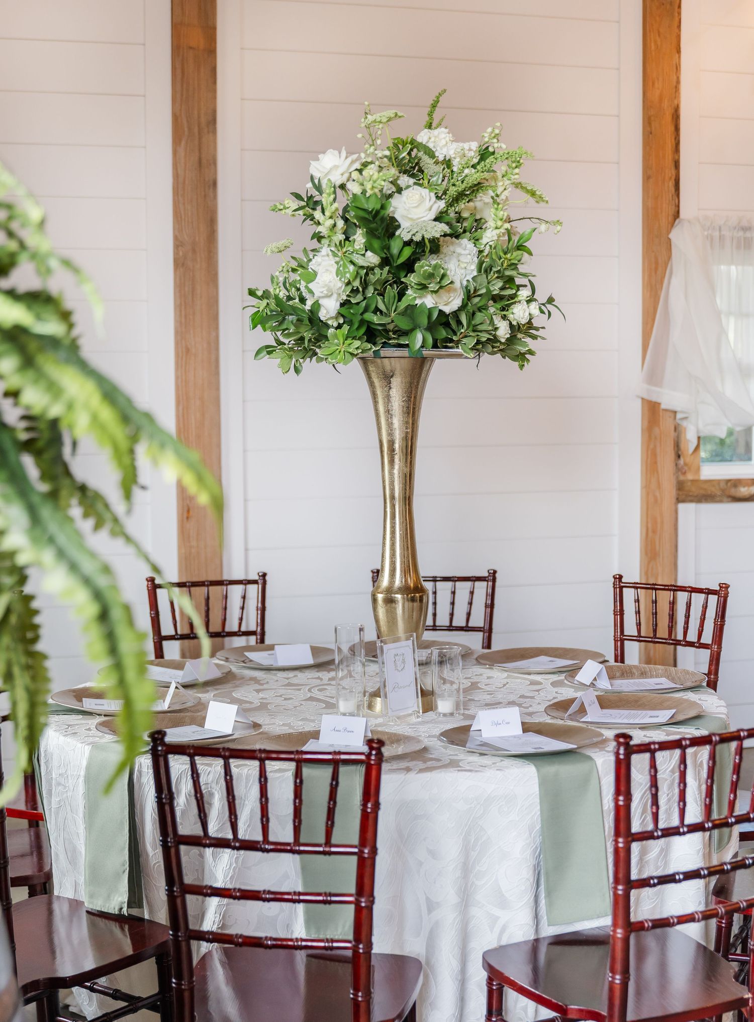 Elegant wedding reception tables set with white linens, mahogany Chiavari chairs and tall floral centerpieces in gold vases.