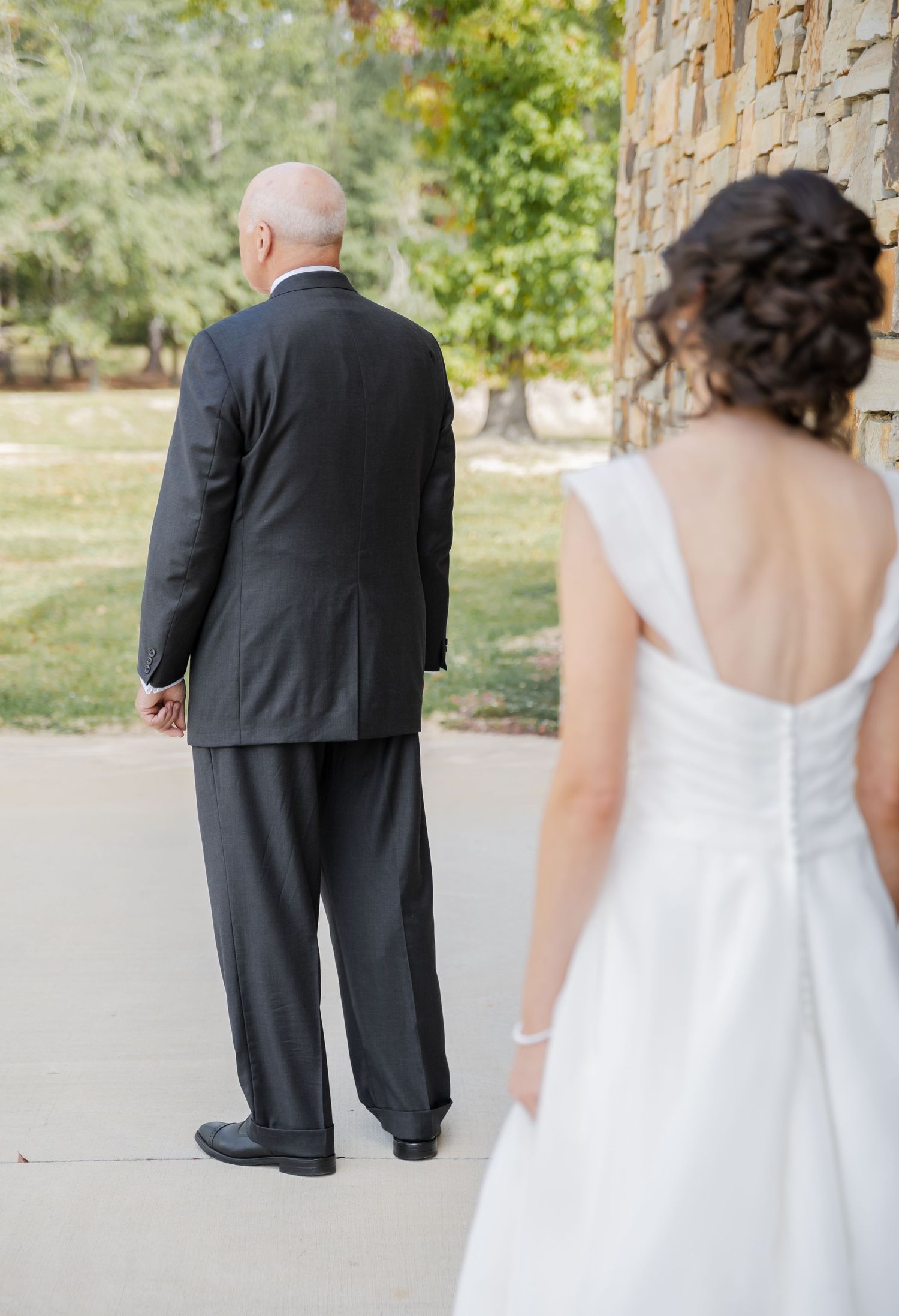 A bride in a white dress stands behind someone wearing a black suit at an outdoor wedding venue.