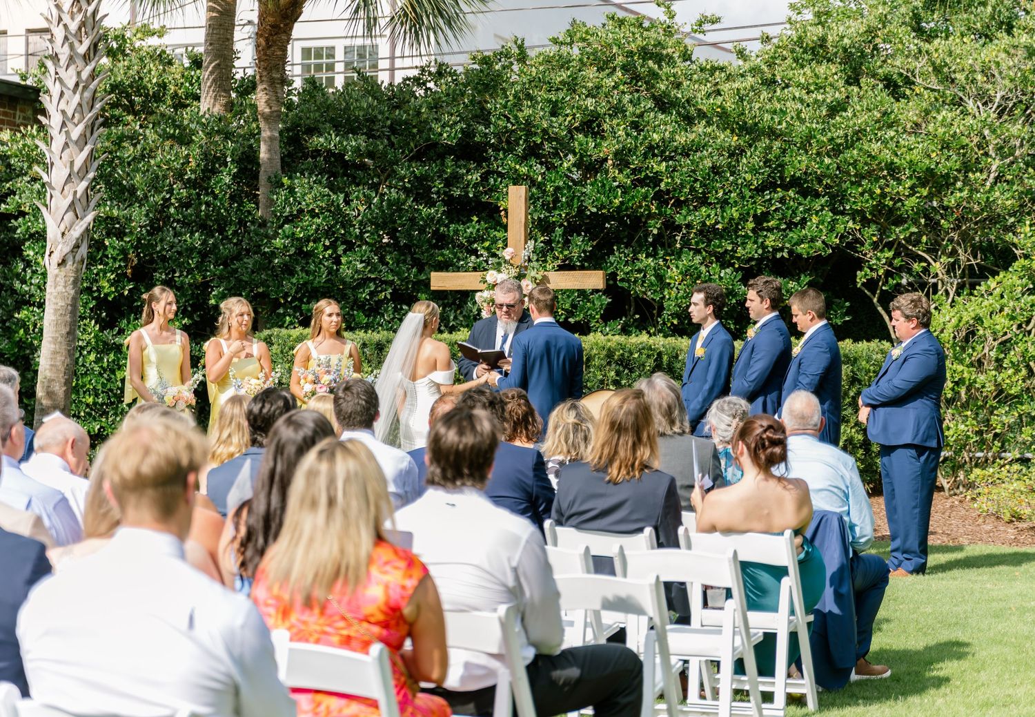Guests seated in white chairs for an outdoor garden wedding ceremony with palm trees and greenery in the background.