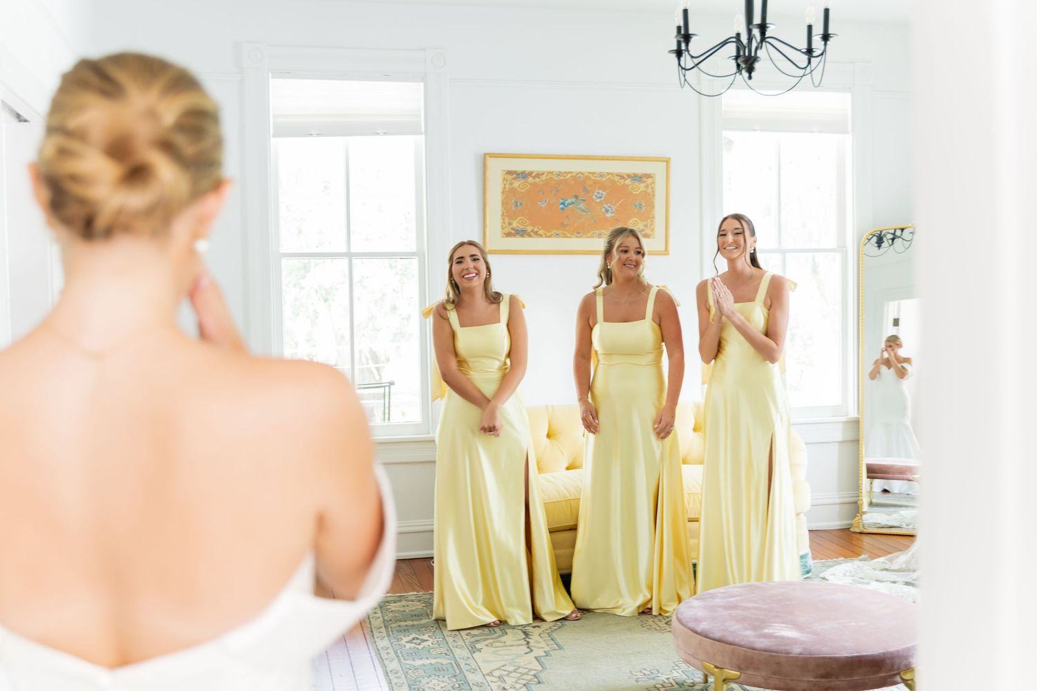 Bridesmaids in pale yellow dresses react with joy as they see the bride for the first time in a bright white room.