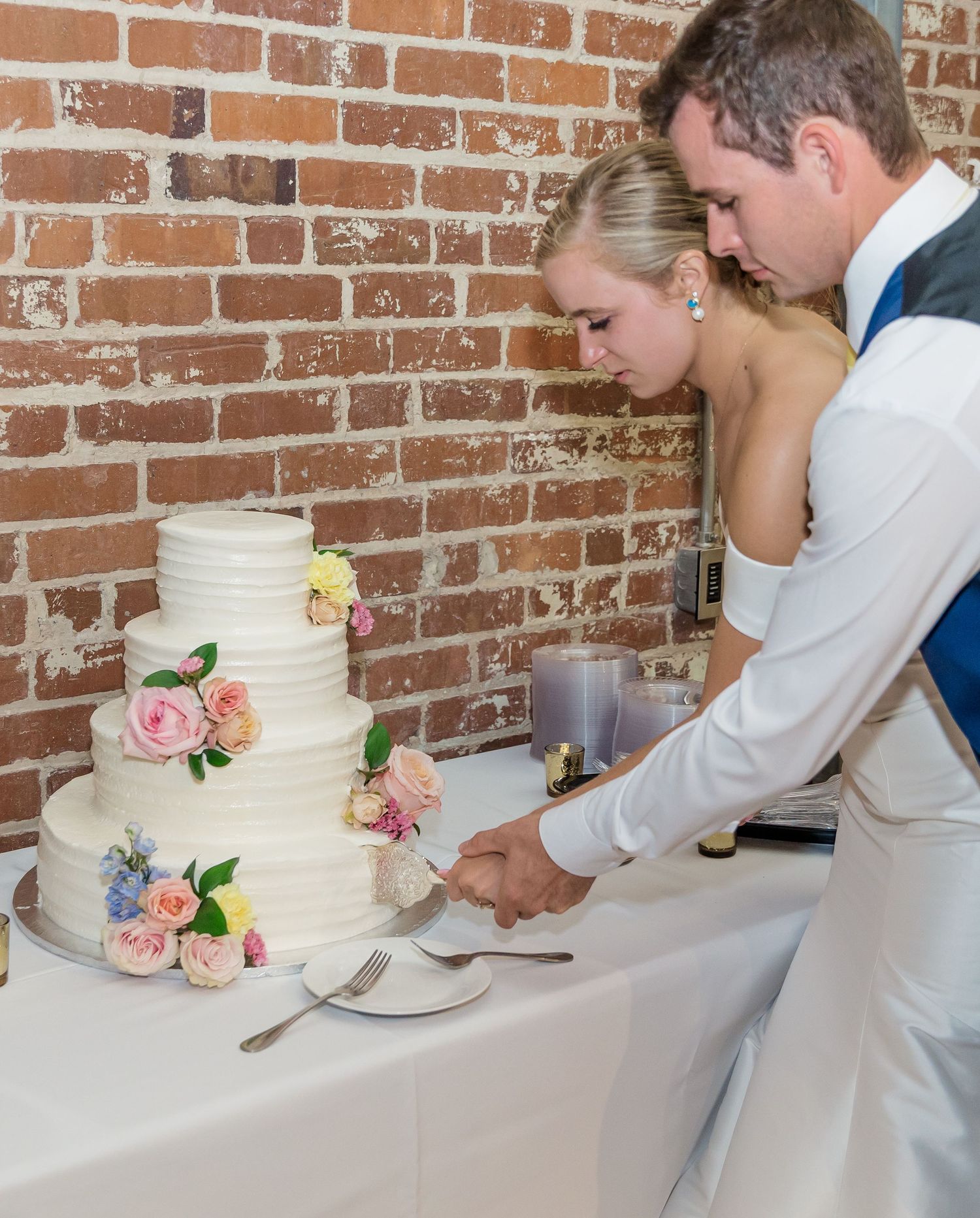 Newlyweds perform traditional cake cutting ceremony at their wedding reception against a rustic brick wall backdrop.