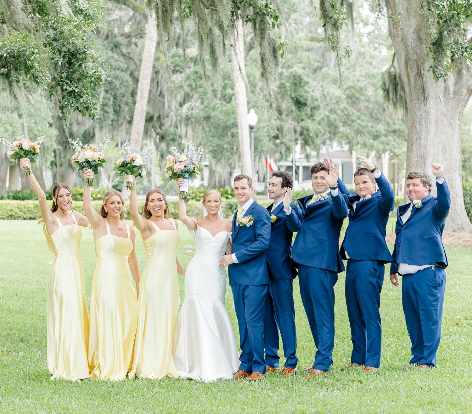 Wedding party in navy blue and yellow attire poses together on a tree-lined lawn during outdoor celebration.