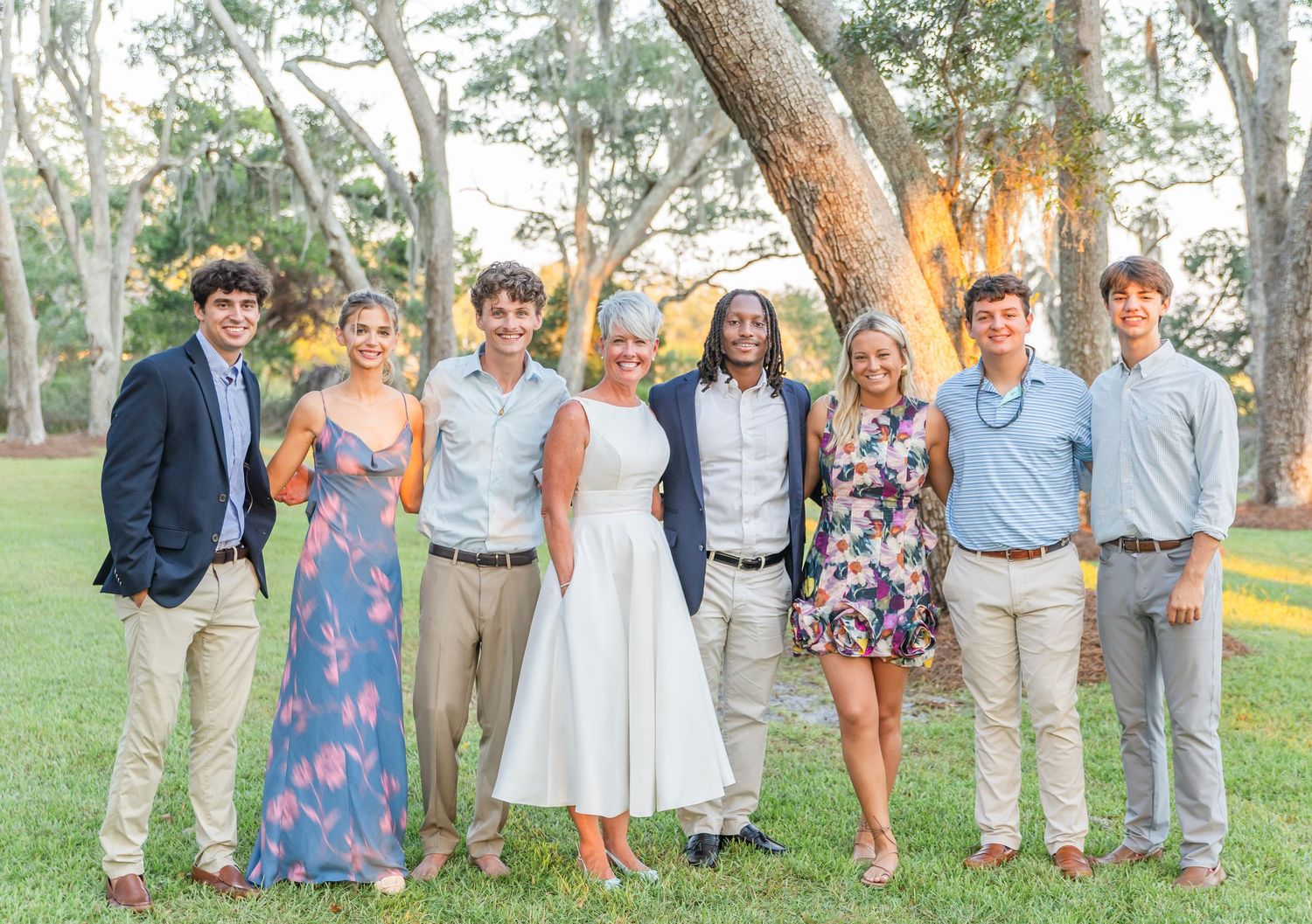 Group in formal attire poses together on a grassy lawn with trees in the background during a summer event.