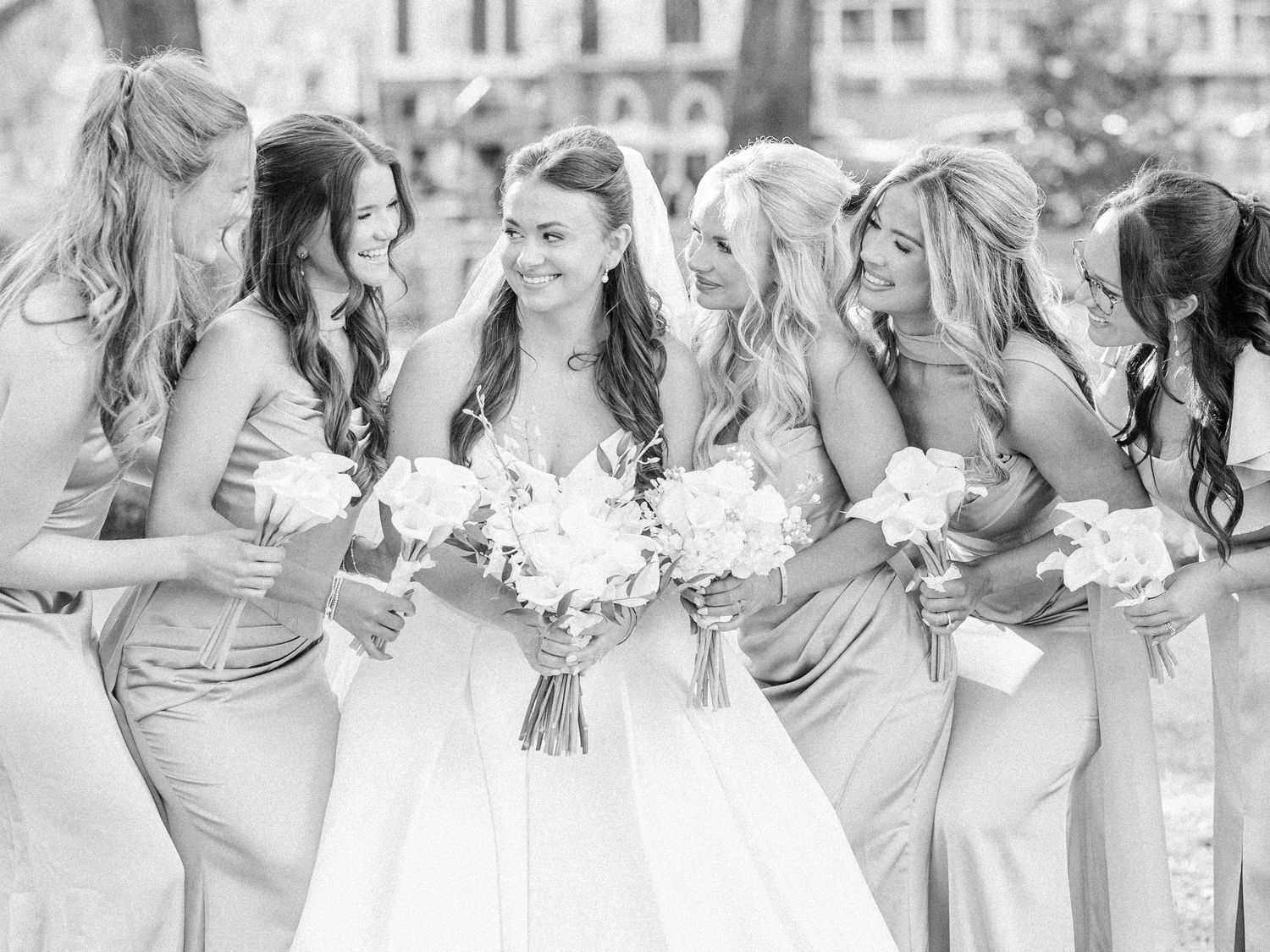 Wedding party in gray dresses laughs together while holding white floral bouquets outdoors.