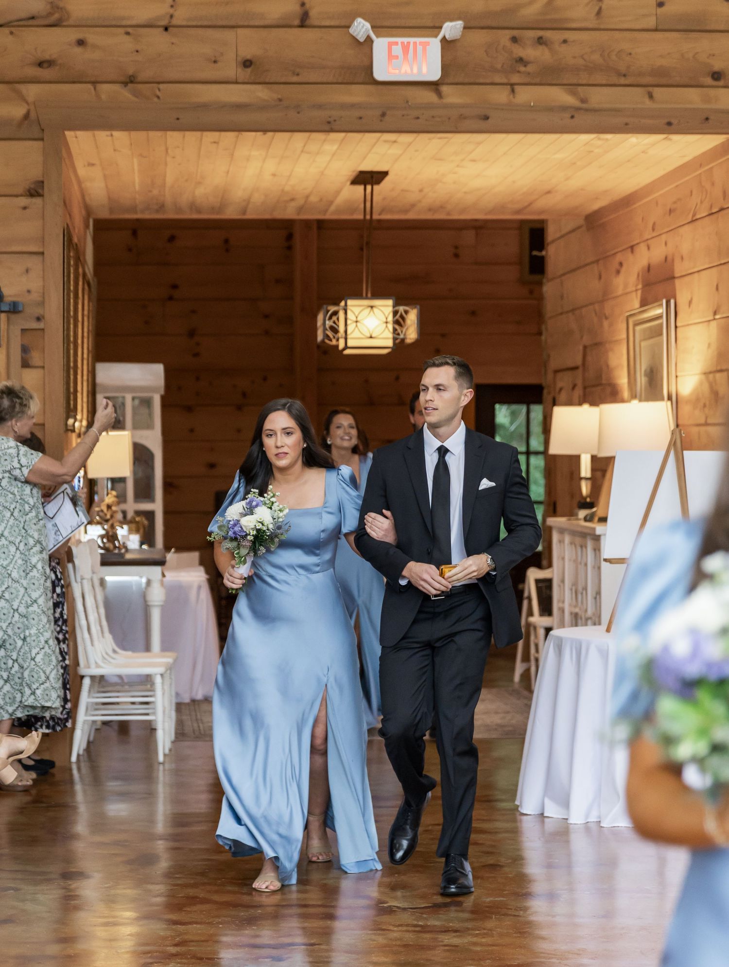 Wedding party members in light blue dresses and dark suits dance together in a rustic wooden reception venue.