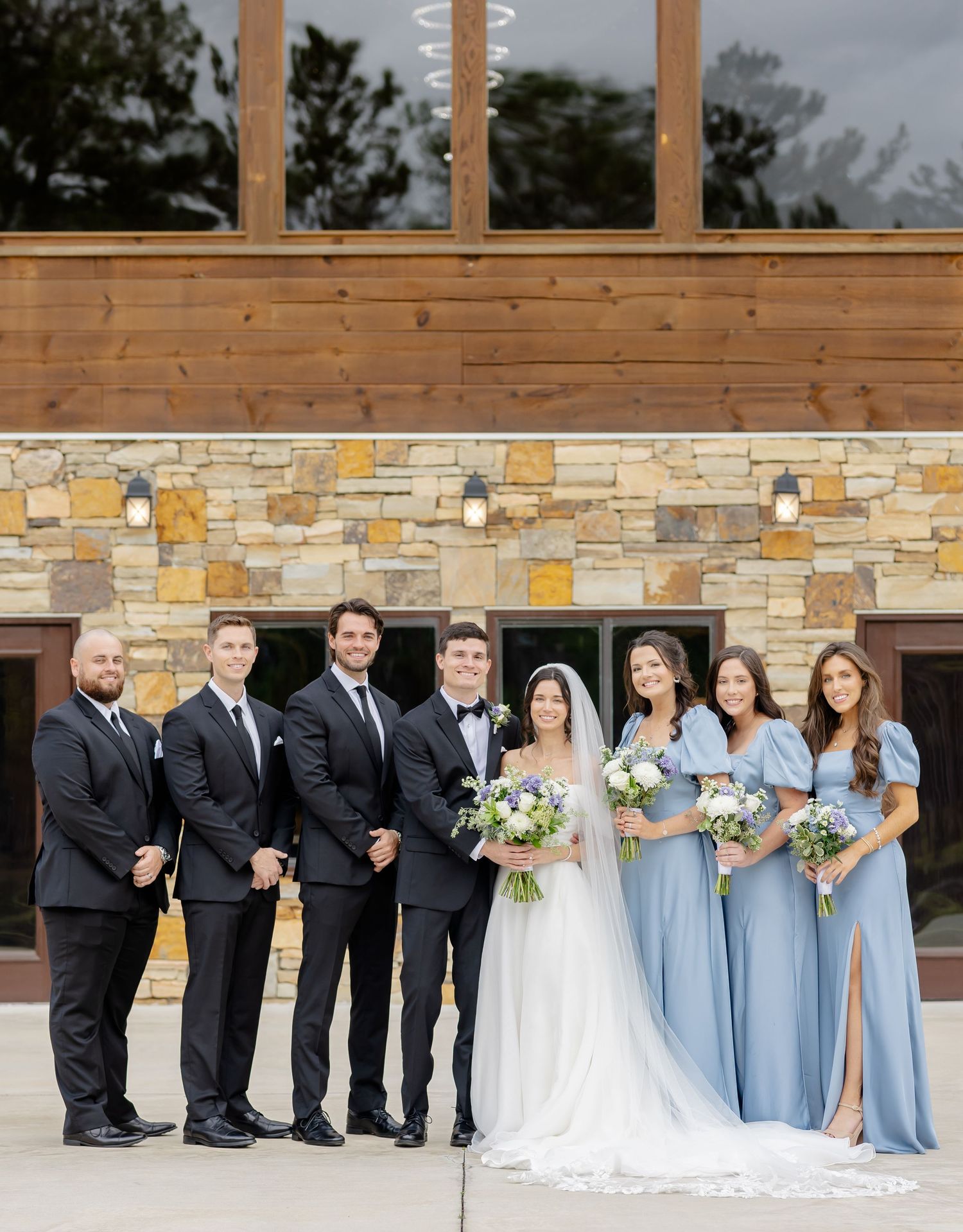 Wedding party in formal attire poses against stone and wood wall, with bridesmaids in light blue dresses.