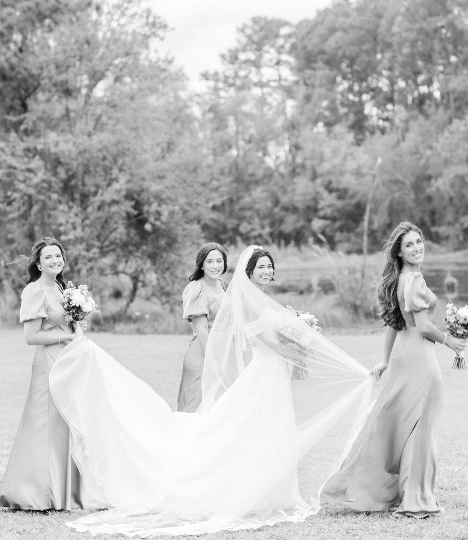 Bridesmaids in light colored dresses pose with flowing wedding gowns in a scenic outdoor setting.
