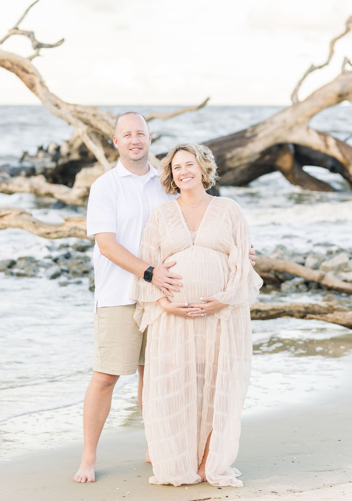 Expectant couple poses on beach with driftwood, wearing light colored casual attire.