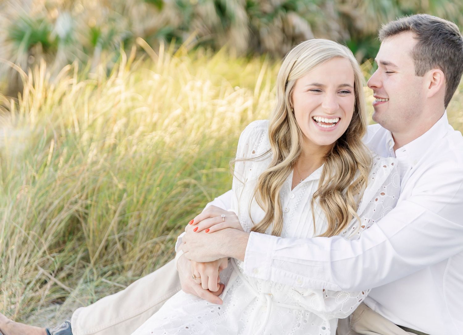 A couple wearing white outfits shares a joyful moment together while sitting in tall golden grass.
